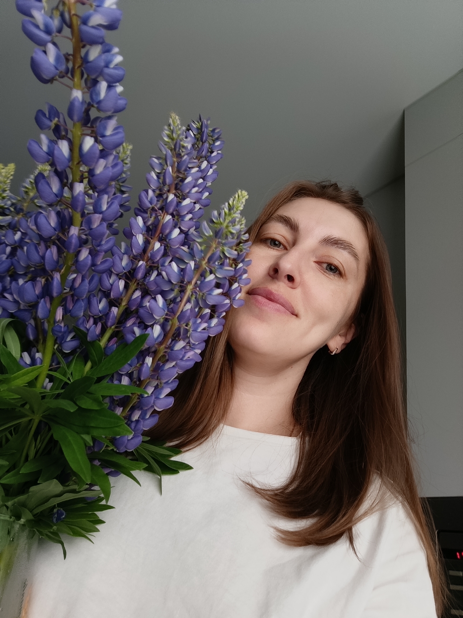 A woman with long, light brown hair is holding a bouquet of purple lupine flowers. She is wearing a white t-shirt and has a small earring in her right ear. The background appears to be a room with grey walls and a portion of a cabinet or appliance visible in the lower right corner. The location is Chaikovsky, Russia.