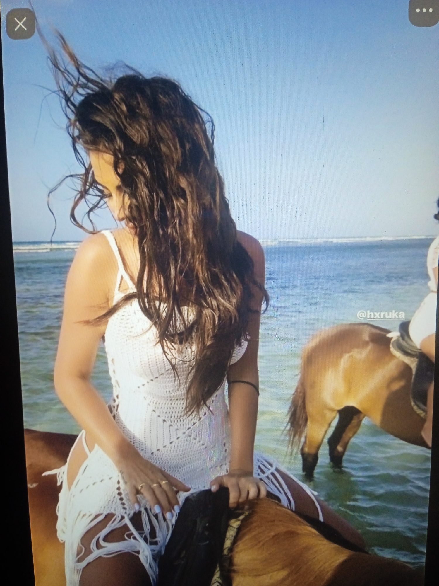 The image shows a woman with long, dark, wet hair riding a horse in shallow water. She is wearing a white crocheted dress with fringe. The horse is brown. The water appears to be the ocean, with waves visible in the background under a clear blue sky. A social media handle "@hruka" is visible on the right side of the image. There is no information in the image to suggest this location is in Chernivtsi, Ukraine.