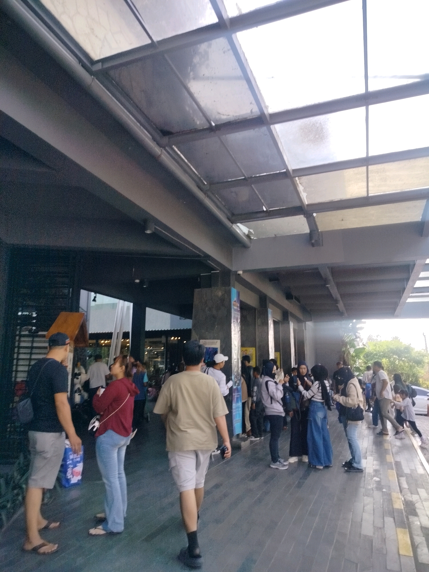 A group of people stand on a paved walkway under a covered outdoor area with a translucent roof. The walkway is made of large, grey rectangular tiles with yellow accents. The covered area has a dark grey, angled ceiling structure with multiple transparent panels that allow light to filter through. In the background, more people are visible, some walking and others standing in groups. There are also glimpses of greenery and buildings.
On the left side of the image, three individuals are most prominent. A man wearing a dark t-shirt and grey shorts stands with his back to the camera, holding a blue shopping bag. To his right, a woman in a red hooded jacket and blue jeans faces away from the camera, looking to her right. Another man in a light brown t-shirt and light-colored shorts is walking away from the camera, facing right.
Further back, to the right of the main group, several other people are gathered. Some are wearing hijabs and casual clothing, standing and talking. A white car is visible in the far background on the right. The overall scene suggests a public area, possibly an entrance or walkway of a commercial or recreational facility in Megamendung, Indonesia.
