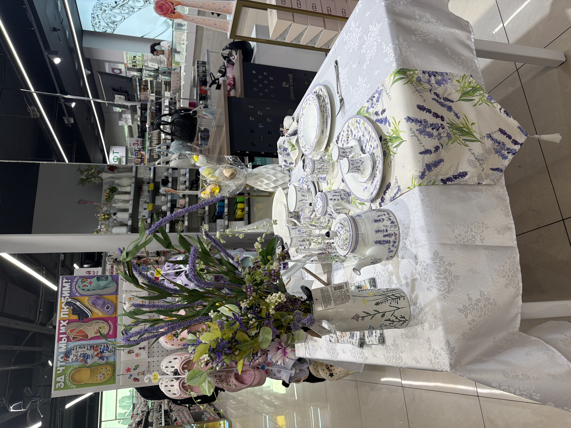 The image shows a close-up of a table setting with a floral theme, likely in a retail store in Sochi, Russia. A white tablecloth with a subtle embossed floral pattern covers the table, and a lavender-themed table runner is laid across it. On the table, there is a white teapot with a lavender design, matching plates, teacups, and saucers, as well as some glassware. A vase filled with a bouquet of lavender and other flowers is also present, adding to the decorative display.
In the background, the scene suggests a store environment. Shelves with various merchandise, including bags and what appear to be decorative items or homeware, are visible. A sign in Russian, displaying images of colorful clogs and the text "ЗА ЧТО МЫ ИХ ЛЮБИМ?" (Why do we love them?), is prominently displayed on the left side of the image, hinting at a shoe or clothing store. Further back, a person can be seen in the background, possibly a store employee. The lighting suggests it is daytime, and the indoor setting implies the weather is not a factor. The overall impression is a display designed to be attractive and inviting, showcasing a coordinated set of tableware and decorative items. The presence of a Pierre Cardin logo on a box in the background further contextualizes the setting as a retail space.