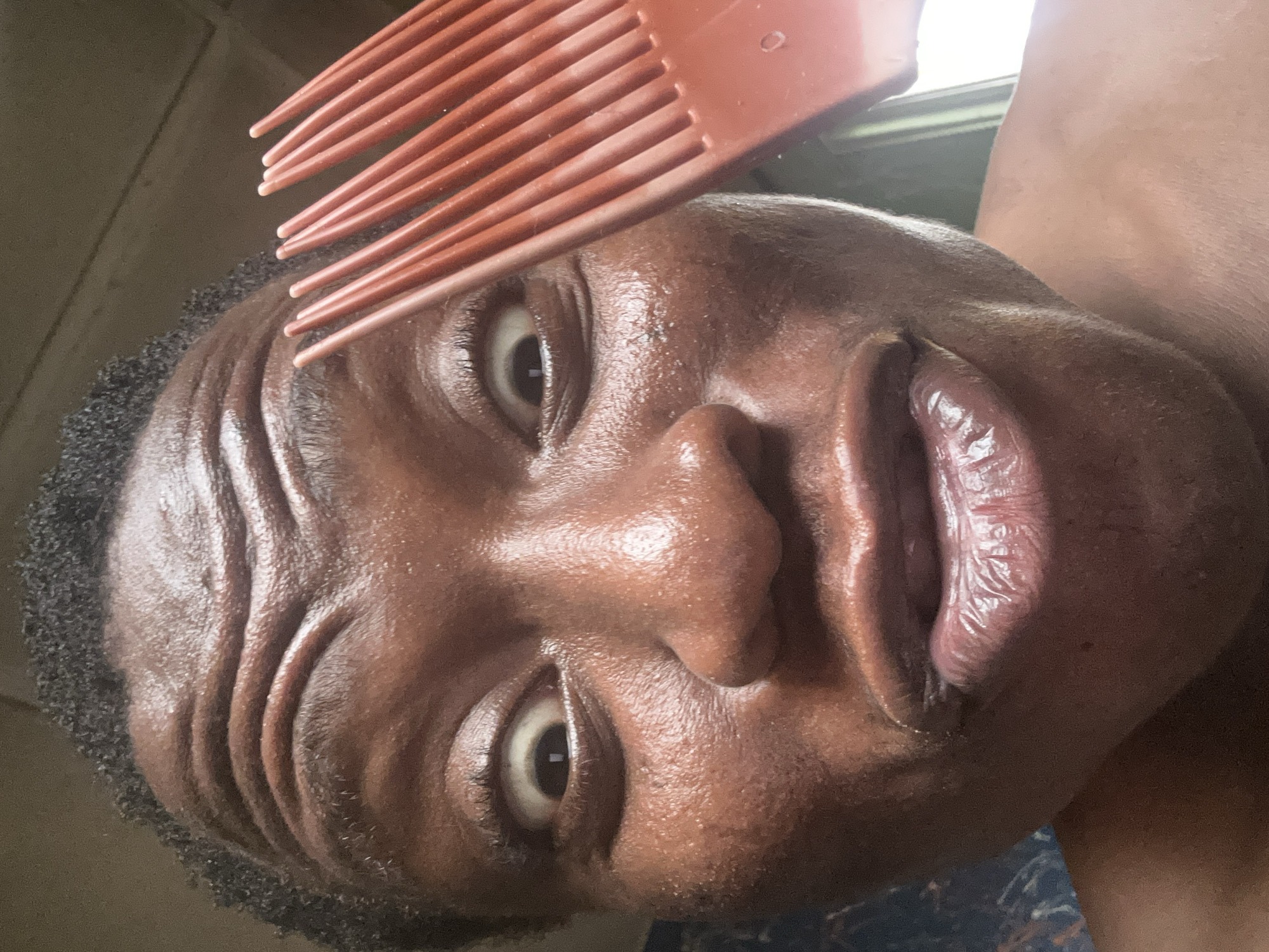A close-up, rotated image features a person with dark skin, likely male, looking directly at the camera with a wide-eyed, expressive facial expression. An orange or reddish-brown afro pick is positioned over the top of their head, with its teeth partially visible, suggesting it's either in the process of being used for hair styling or playfully placed.
The person's face shows notable details: the skin appears shiny or oily, reflecting light, particularly on the forehead, nose, and cheeks. Deep creases are visible on the forehead, likely caused by the upward movement of the eyebrows, contributing to the expressive look. Their eyes are wide open, revealing significant whites around the dark pupils. The lips are pursed or puckered, adding to the exaggerated expression. Short, dark, curly hair frames the face. The background is indistinct and dark, with some brighter areas suggesting a light source or window to the upper right. No text is visible in the image. The time of day, weather, and specific location cues are not discernible from the provided media. The overall emotion conveyed is one of surprise or playfulness due to the facial expression.