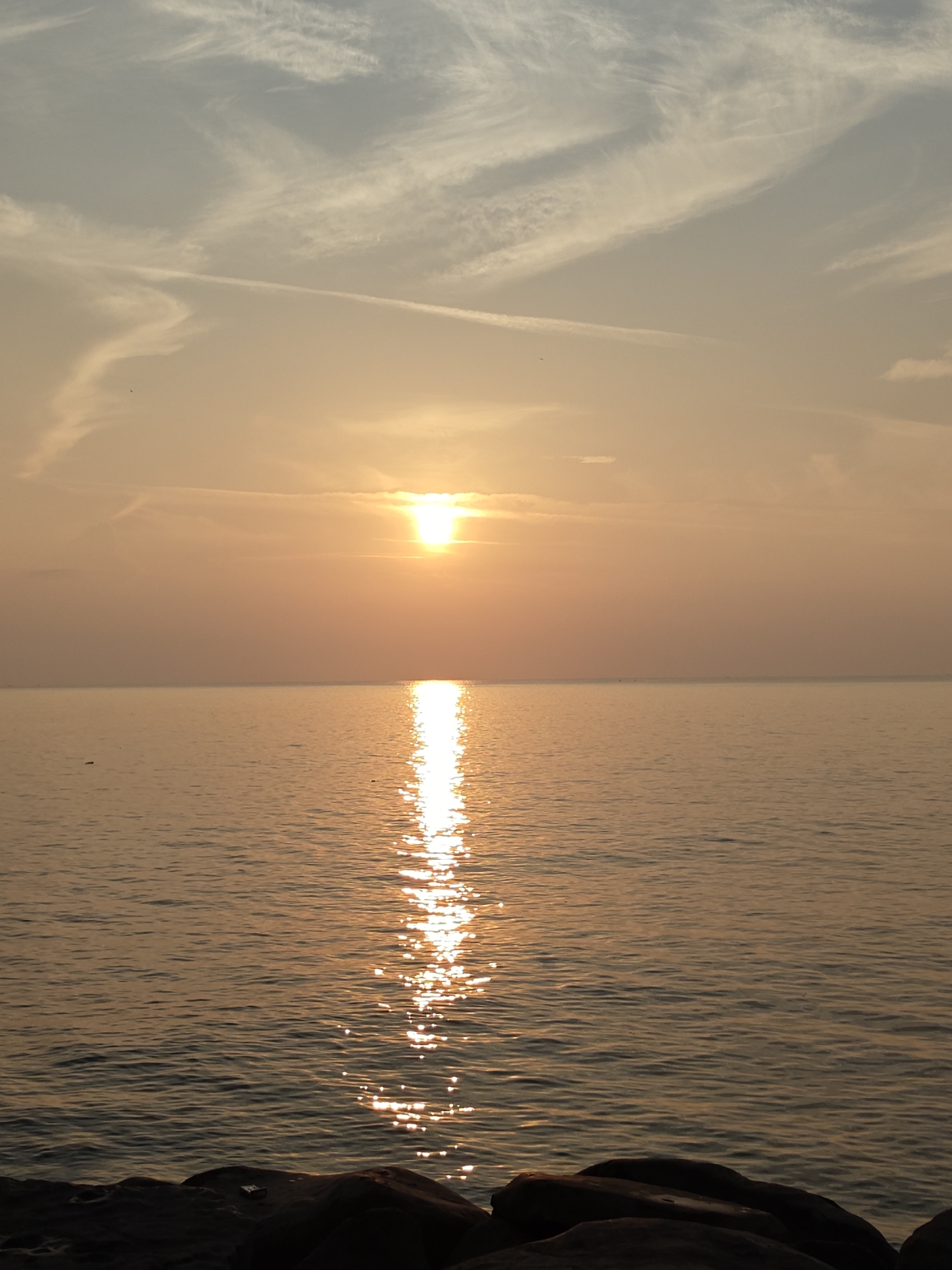 The image depicts a serene sunset over the waters of Lhokseumawe, Indonesia. The sun, a bright, warm orb, is positioned slightly left of the center of the frame, casting a radiant golden glow across the sky. Delicate, wispy clouds streak across the horizon, tinged with the orange and yellow hues of the setting sun.
Below the sky, the calm sea stretches to the horizon, reflecting the sun's light in a shimmering path that leads directly towards the viewer. The water's surface is gently rippled, creating a textured effect of light and shadow. In the foreground, dark, silhouetted rocks line the edge of the water, providing a grounding element to the vast expanse of sky and sea. The overall impression is one of tranquility and natural beauty.