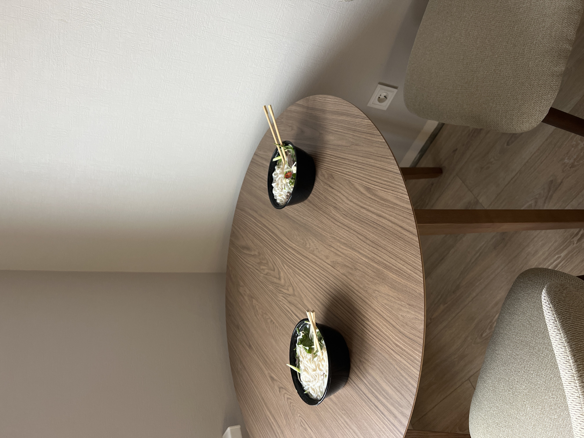 The image shows a high-angle view of a round, wooden table in a room with light-colored walls. Two black bowls of noodles with chopsticks are placed on the table. Two chairs with light-colored, textured upholstery are positioned near the table. The floor appears to be wooden. An electrical outlet is visible on the wall. The image's location is not possible to determine from the given information.