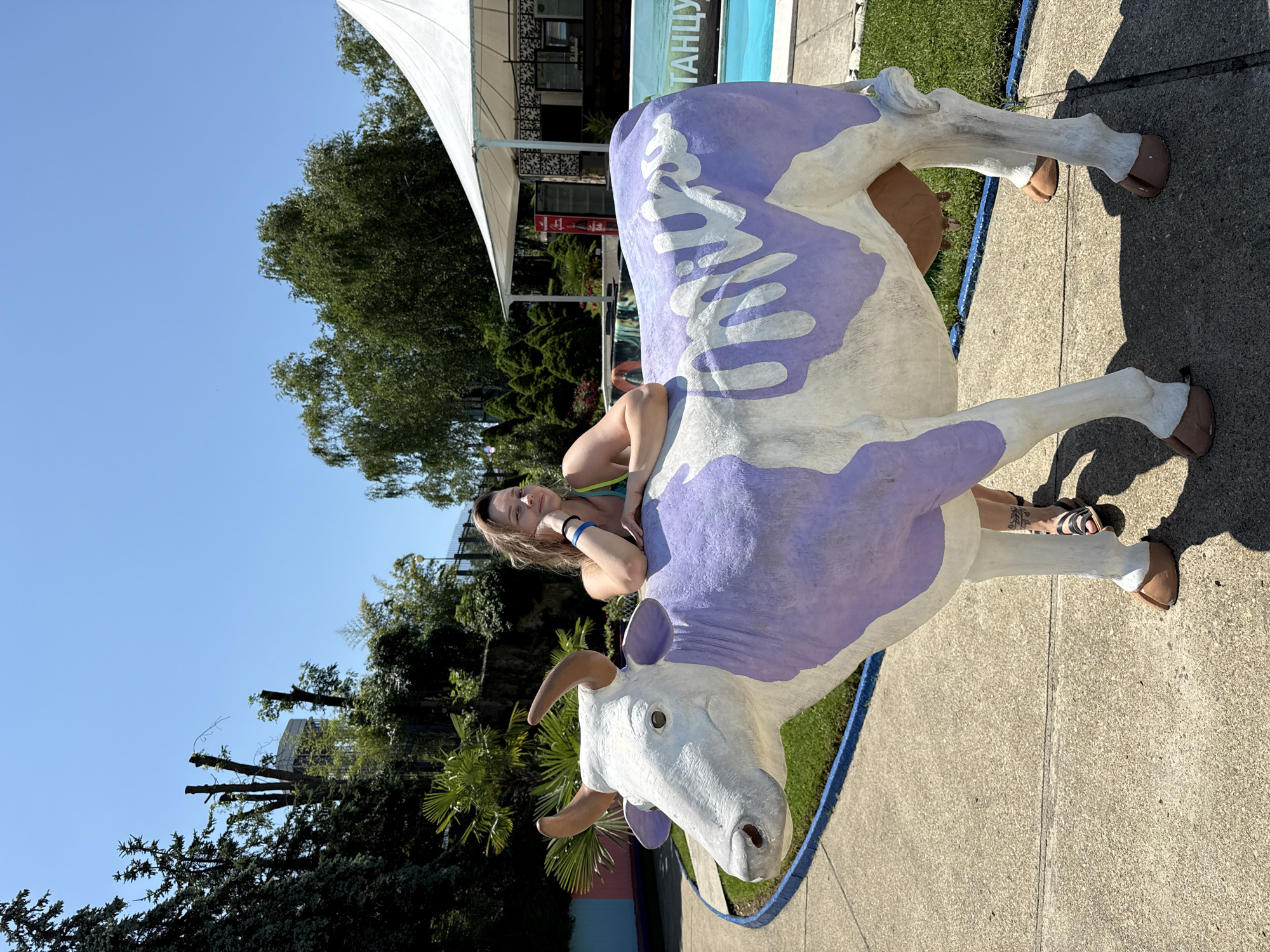 In Nebug, Russia, a woman poses with a large, whimsical statue of a cow painted white with purple splotches. The cow statue is positioned on a paved walkway. The woman, wearing a bright yellow and green bikini top and black sandals, leans on the cow's back, resting her head on her hand. She has a tattoo on her leg. Behind the cow and woman, a swimming pool is visible, with a white tented structure in the background. Lush green trees and bushes surround the area, and the sky is clear blue. A sign with Cyrillic writing, likely indicating a local establishment or attraction, is partially visible near the pool.