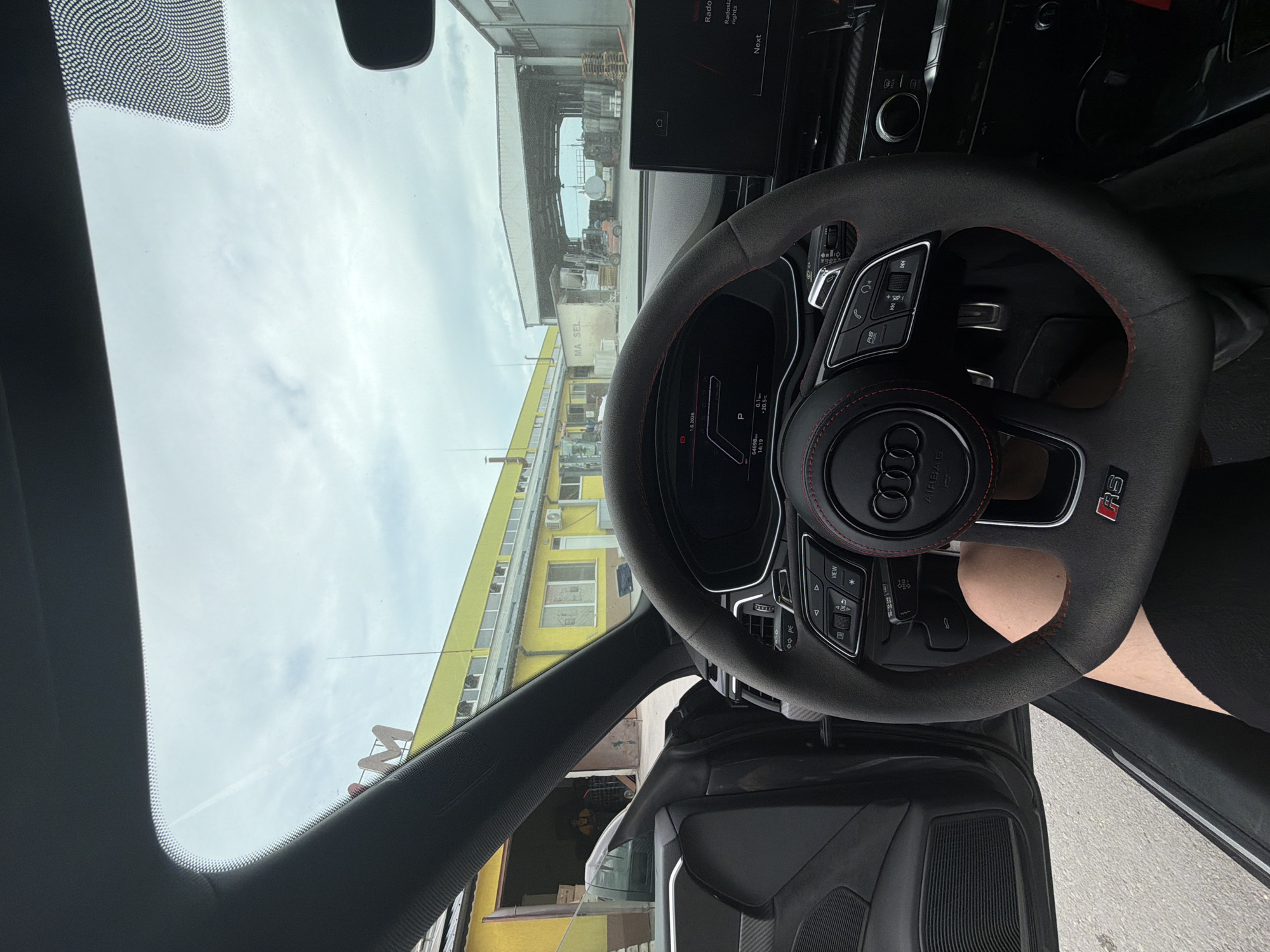 The image, taken from inside a car, shows the dashboard, steering wheel, and the view through the windshield. The steering wheel, with the Audi logo and "RS" emblem, is prominently centered. Through the windshield, a yellow building and a warehouse-like structure are visible, likely an industrial or commercial area. The digital display on the dashboard shows the date and time, along with other car-related information. The location is likely Dolno Ablanovo, Bulgaria, based on the presence of a sign with "MA SEL" on the yellow building, a possible business name. A person's leg is visible resting on the door sill.