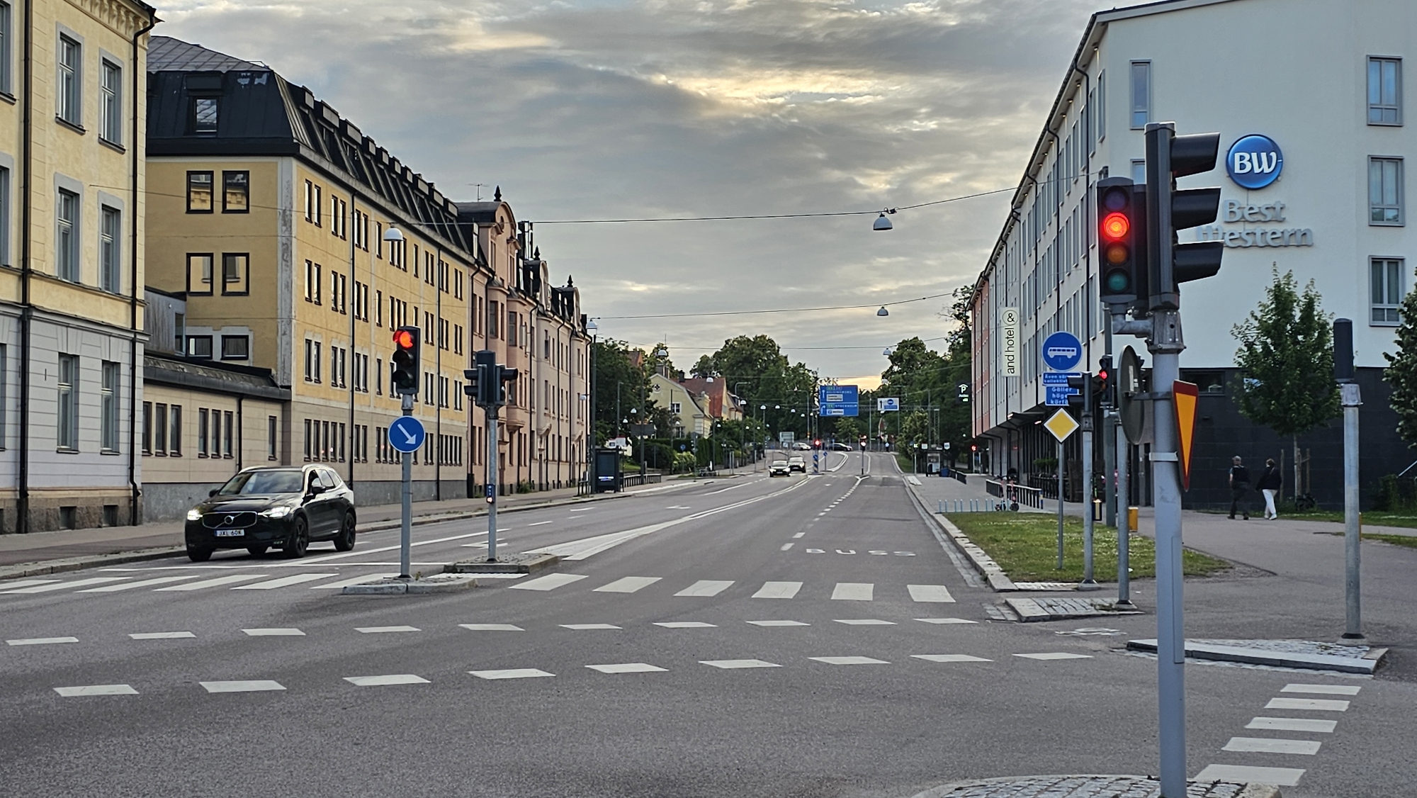 This media file depicts a street scene in Linköping, Sweden, likely during the late afternoon or early evening. The sky is overcast with dramatic clouds, suggesting an approaching storm or the end of the day.
The foreground features a wide asphalt road with clearly marked pedestrian crossings. A black SUV is stopped at a red traffic light on the left side of the road, positioned at a pedestrian crossing. To the right, a modern hotel with a "BW Best Western" sign is visible. A traffic light controlling traffic on the right side of the road is also red.
In the midground, the road stretches into the distance, lined with buildings on both sides. On the left, a row of older, multi-story buildings with yellow facades and dark roofs are visible. On the right, the modern hotel continues, followed by trees and more buildings further down the road. Several cars are driving on the road in the distance, and more traffic lights are visible, some showing red.
Notable details include street lamps, power lines, and various road signs, including a blue sign with white text and arrows indicating directions, and a bus stop sign. Two people are walking on the sidewalk in front of the hotel on the right. The overall atmosphere is one of urban normalcy, with traffic and pedestrians coexisting under a dramatic sky. The presence of a "BUS" marking on the road indicates public transportation routes.