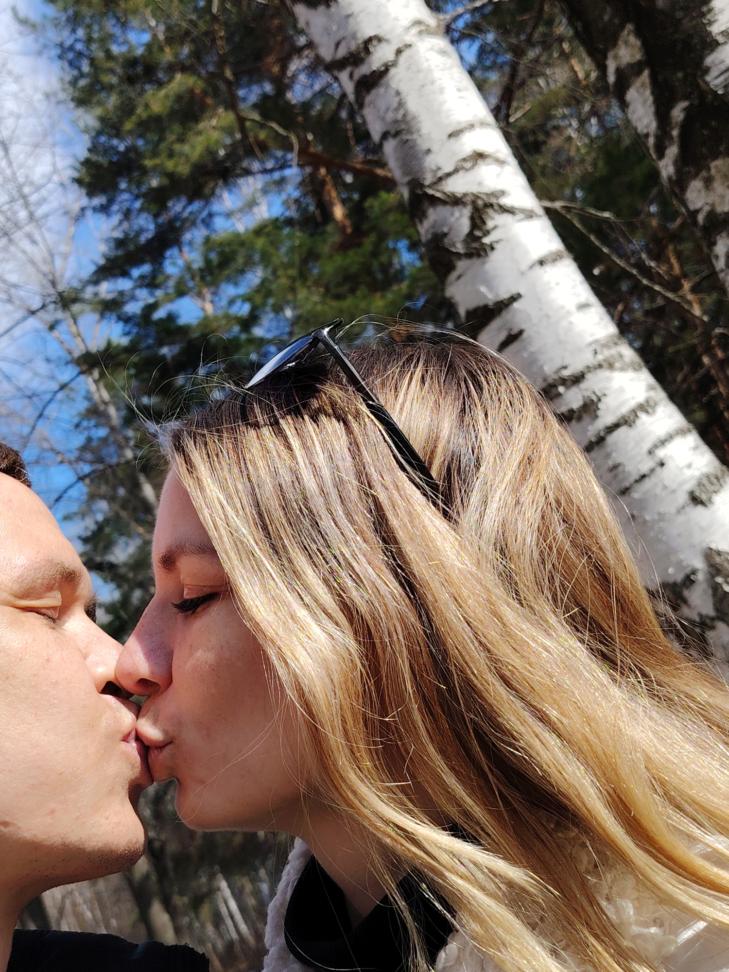 In the city of Chaikovsky, Russia, a couple is intimately kissing. The female has long, blonde hair with sunglasses perched on top of her head. The background includes a birch tree and glimpses of blue sky and green trees.