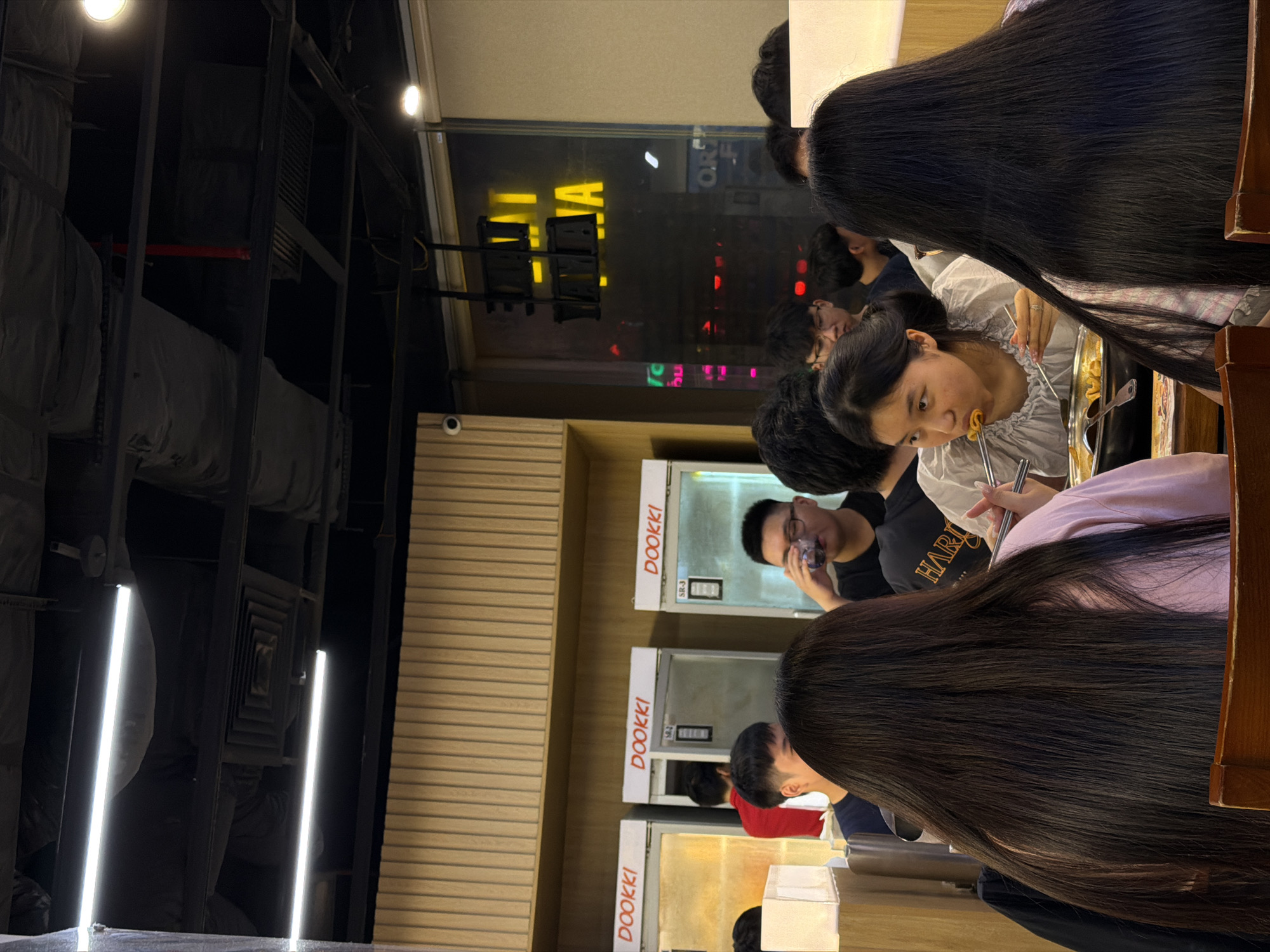 The image shows a group of young people inside a restaurant, likely a Korean tteokbokki restaurant given the visible "DOOKKI" text on the service counter. The scene is brightly lit, suggesting it's daytime or a well-lit interior. Most of the people are facing away from the camera, engrossed in their meal. One young woman in the foreground is actively eating, using chopsticks to pick up food from a communal pot. Her expression appears focused and engaged with her food.
The restaurant's interior features wooden paneling and metal shelving, creating a modern and functional aesthetic. In the background, through a large window, city lights are visible, hinting at an urban setting. The overall atmosphere seems casual and social, indicative of friends or family enjoying a meal together. The prominent black hair of several individuals suggests an East Asian demographic. There are no clear indicators of the weather or specific time of day beyond the lighting. The visible text "DOOKKI" is a brand name, and "SR-3" and "SR-2" are likely item or table identifiers. The image is captured in portrait orientation with the bottom of the image oriented towards the left side of the frame.