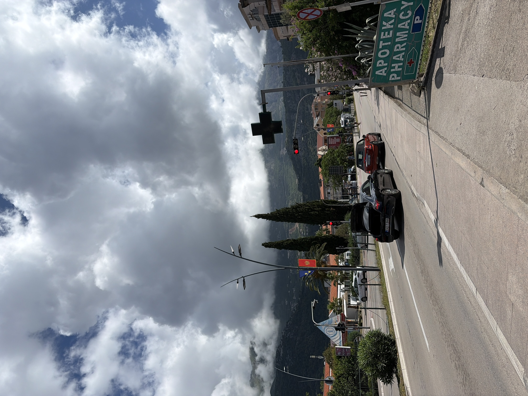 The image, rotated 90 degrees counter-clockwise, depicts a vibrant street scene in Budva, Montenegro, under a partly cloudy sky. The street runs from the bottom left towards the top right, with a paved sidewalk bordering the right side.
In the foreground and midground, several vehicles are present, including a red car with a Serbian license plate "BG 024-KM" and a black car bearing a Montenegrin plate "CG 53-P964". A traffic light, displaying a red signal, is prominently visible over the street. Above the traffic light, a large green cross symbol, commonly associated with pharmacies, is mounted. Further down the sidewalk, a green directional sign reads "APOTEKA PHARMACY" in white text, featuring a smaller green cross and a blue 'P' icon, indicating nearby parking. A red circular prohibitory road sign, with a white background and a red diagonal line, is affixed to a building wall.
The background is dominated by dramatic, green mountains, partially enveloped by white and grey clouds, characteristic of Montenegro's rugged terrain. The street is lined with various trees, including tall, conical evergreens and other leafy species. Flags, specifically the Montenegrin national flag and a blue flag (possibly the European Union flag), are displayed on a pole. Other partially visible signs advertise "EXCURSIONS", "INFO", "MONTENEGRO", "INFORM", "RENT-A-CAR", and "APARTMANI". The scene is captured during daytime, with a mix of direct sunlight and cloud cover.