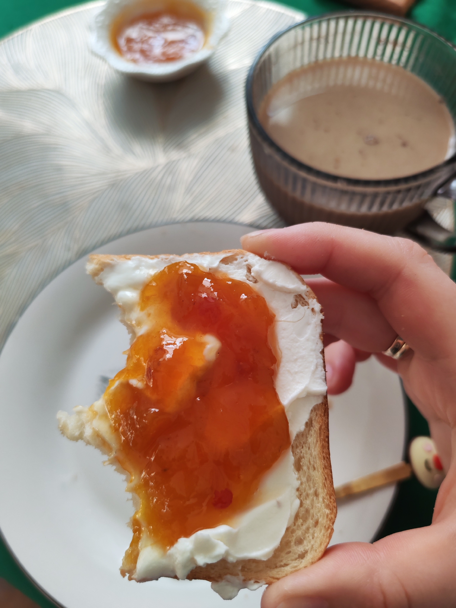 The image depicts a close-up of a slice of bread with cream cheese and orange jam, held by a person's hand. The bread appears to be a whole wheat or multi-grain loaf, with a light brown crust and a soft interior. The cream cheese is spread generously on top, and the jam, which seems to be apricot or peach, is layered over the cream cheese. A small bite has been taken out of the bread.
In the background, a small white dish holds more of the orange jam. To the right, a glass mug filled with a light brown beverage, possibly coffee or tea with milk, is visible. The objects are placed on a patterned placemat with a light, wavy design. A small decorative toothpick with a yellow cartoon-like figure on top is also present.
The scene suggests a breakfast or snack setting. There is no explicit information in the image that would allow for a precise location in Izhevsk, Russia. The image is a still photograph and does not contain any temporal or geographical indicators beyond the visual elements.
