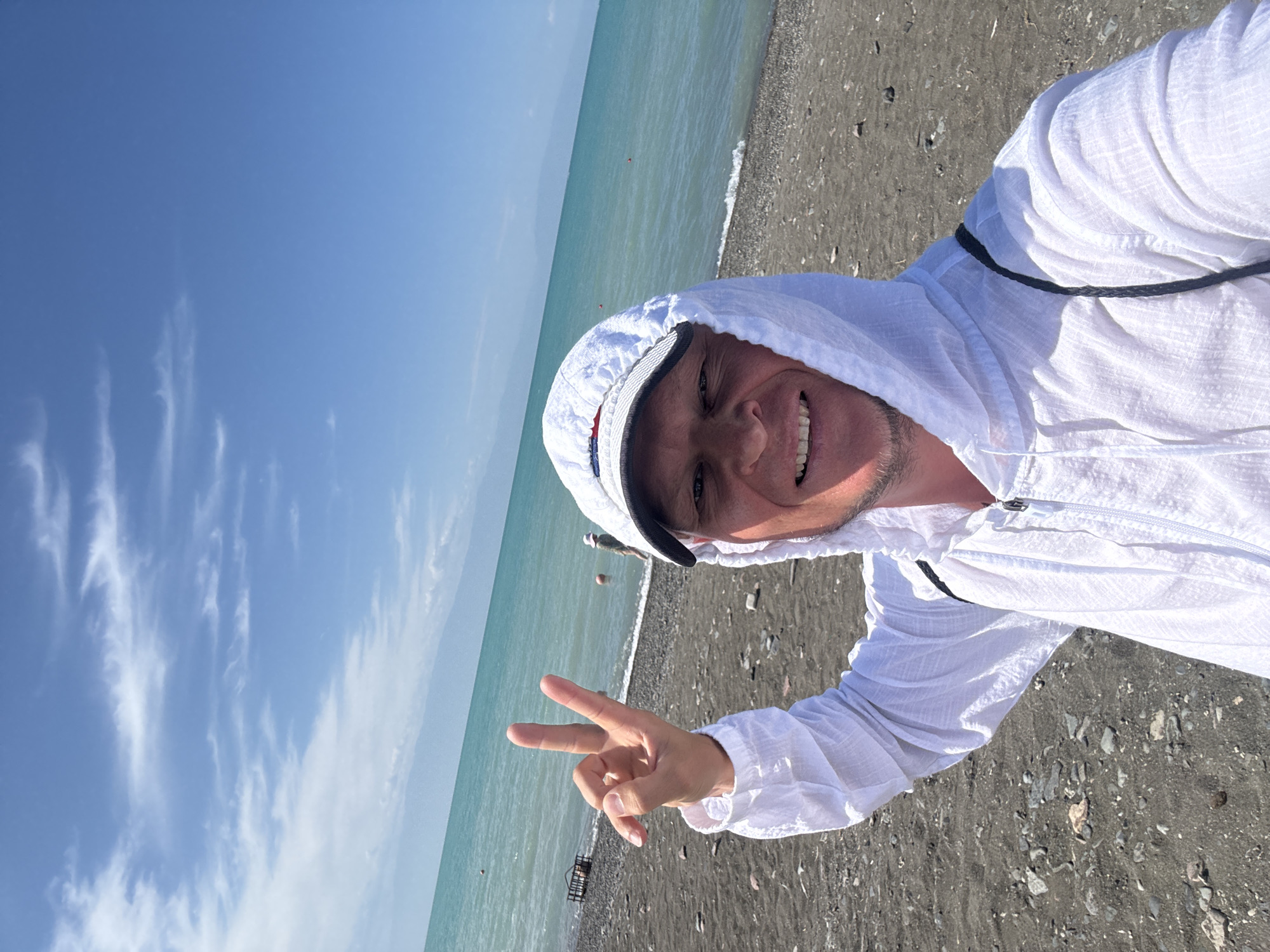 A man with short blonde hair and a light beard smiles at the camera, holding up two fingers in a peace sign with his left hand. He is wearing a white hooded jacket and a white baseball cap with a striped brim. The man is standing on a dark, rocky beach with the turquoise water of a lake behind him. In the distance, faint mountains are visible. The sky is blue with scattered white clouds.