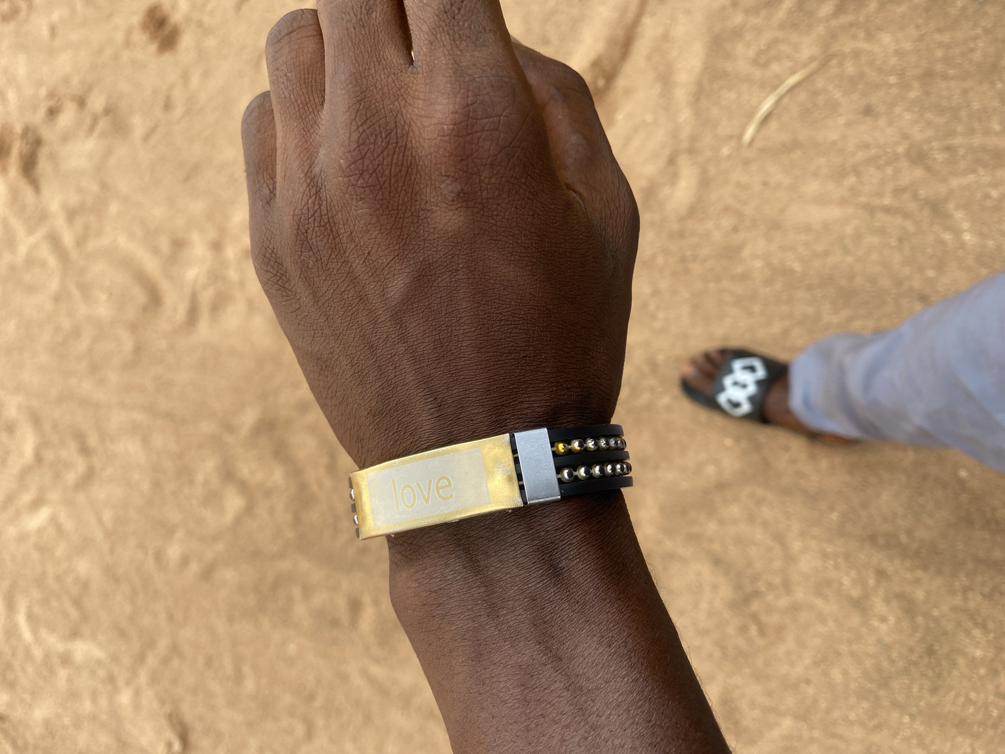 A close-up shot shows a person's left wrist with a bracelet. The bracelet is made of black bands adorned with small, metallic beads and a central gold-toned plate. The plate has the word "love" inscribed on it in a light-colored font. The background is a blurry expanse of sandy ground, suggesting an outdoor setting. In the lower right corner of the image, a portion of another person's leg and foot wearing a light-colored garment and black sandals is visible. The lighting suggests it could be daytime. The overall mood is casual and personal.