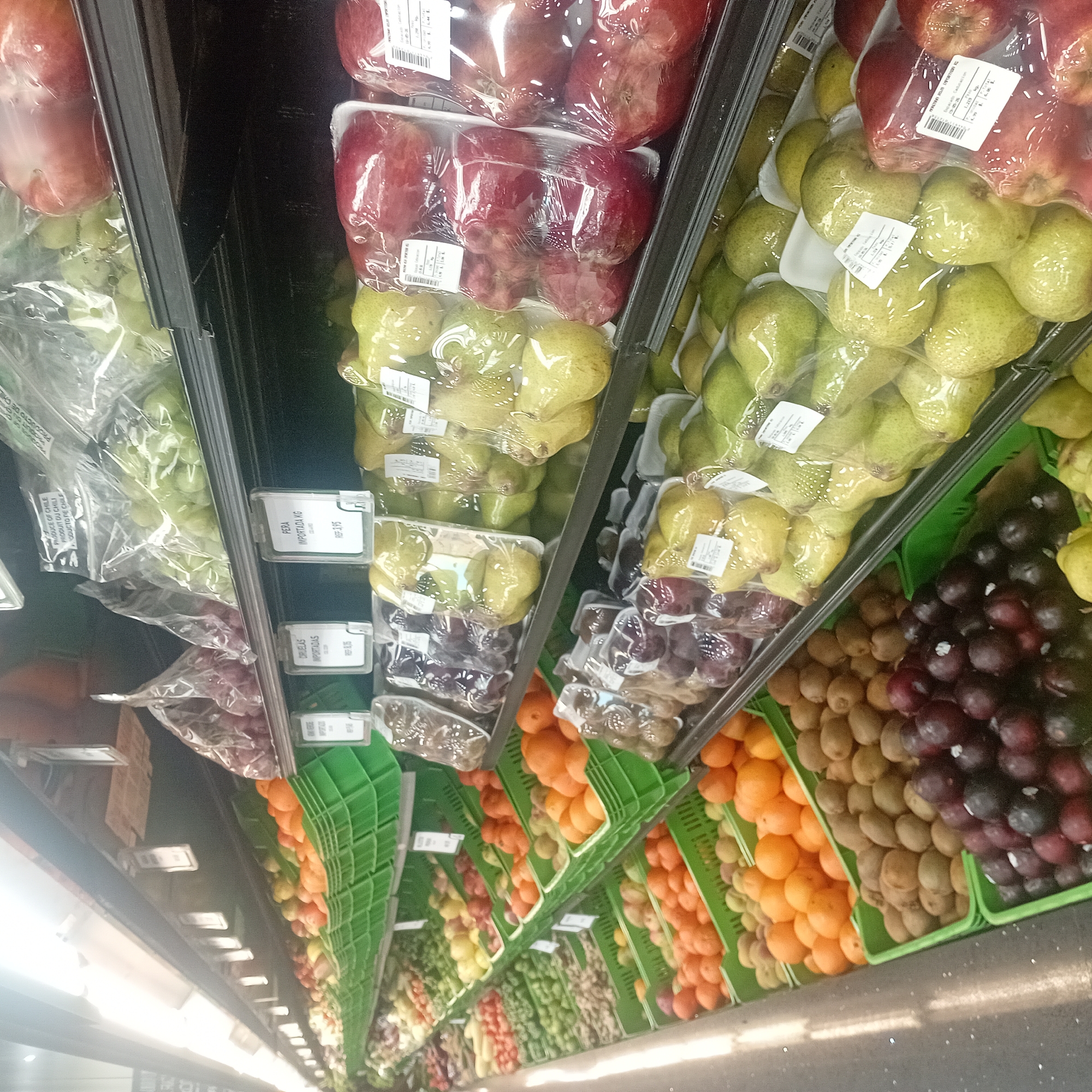 This image shows an aisle in a grocery store. The shelves are stocked with various fruits, packaged and unpackaged. On the top shelves, packaged apples and pears are visible. Below, there are packaged grapes and plums. The bottom shelves contain oranges, kiwis, and grapes in green crates. The fruits are arranged in rows. Price tags are visible on some of the shelves. The image seems to be taken inside a brightly lit store with a black and white speckled floor. The labels indicate that some of the produce is imported.