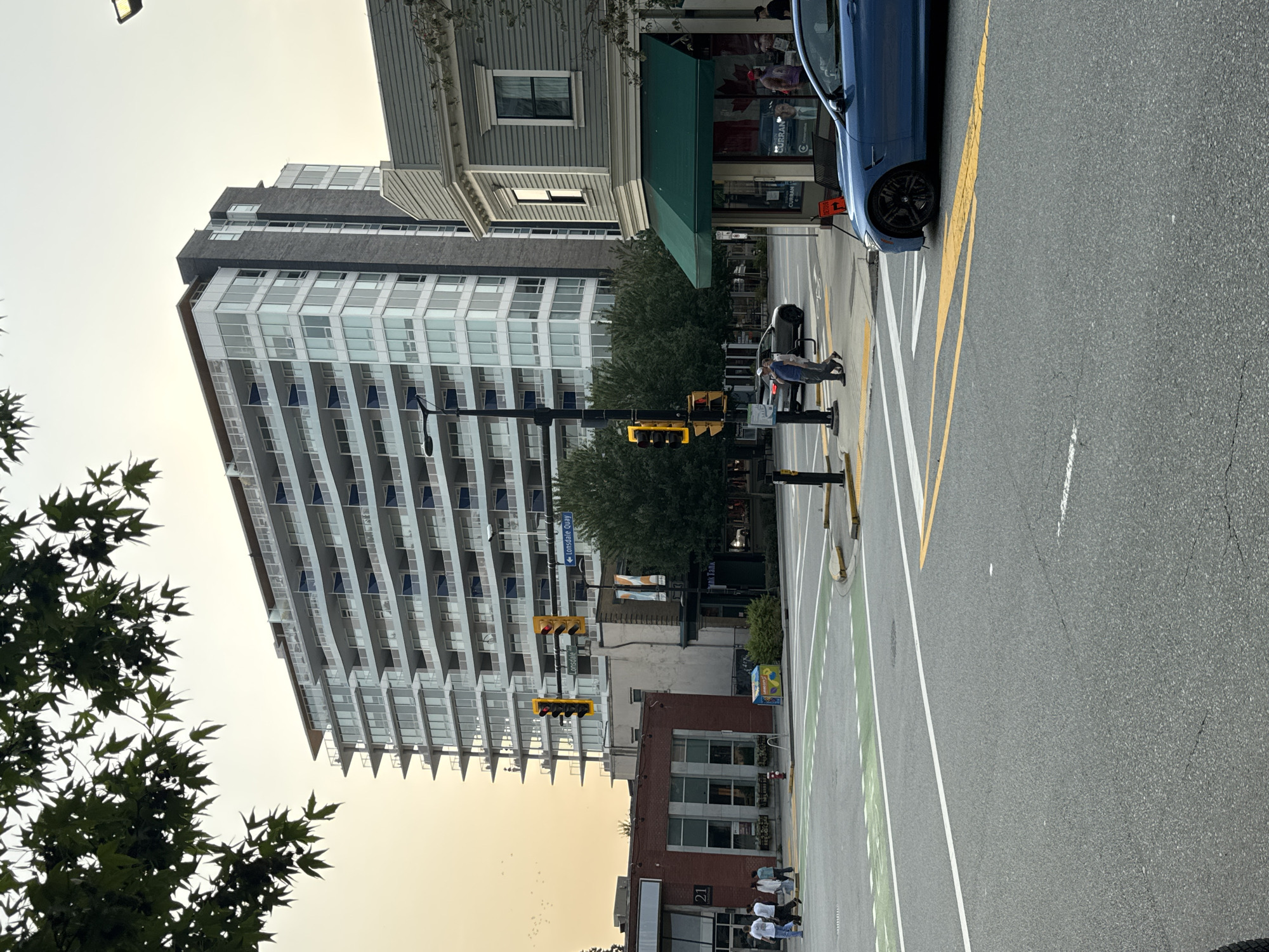 The image depicts an urban street scene in North Vancouver, Canada, likely near Lonsdale Quay given the street signs. A tall, modern apartment building with numerous balconies dominates the background. In the foreground, a blue sports car is parked at an intersection with traffic lights and crosswalks. Pedestrians are visible on the sidewalks and crossing the street. The overall atmosphere suggests a late afternoon or early evening setting with overcast skies. Several trees frame the top of the image, and other buildings, including a brick structure and a grey building with a green awning, are also present. The street is paved and marked with white and yellow lines, indicating traffic lanes and pedestrian areas.