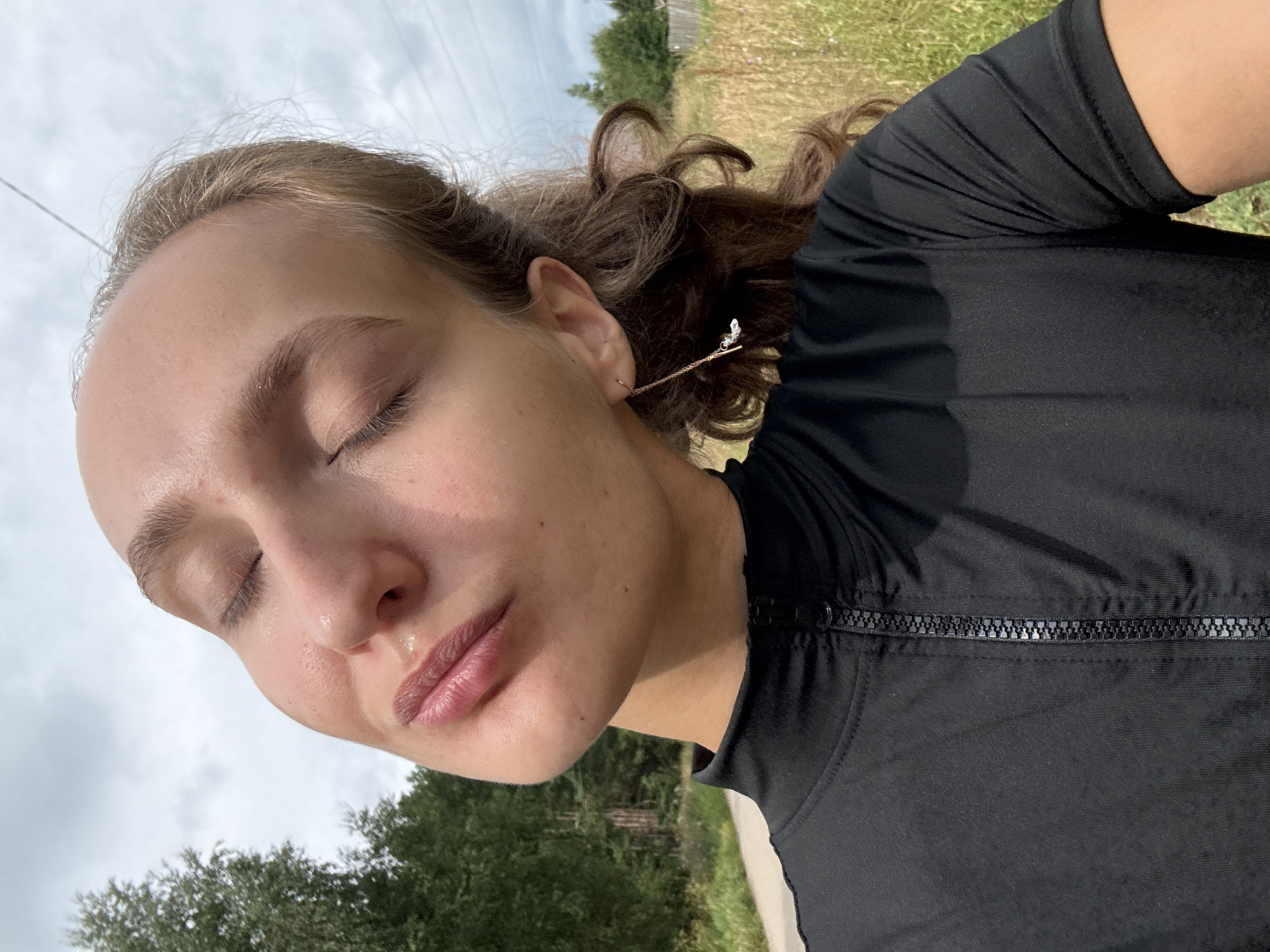 A young woman with blonde hair and closed eyes is taking a selfie outdoors in a rural area, likely the village of Podshivalovo, Russia. She is wearing a black zipped-up top and a long, delicate gold earring with a small charm. Her face is tilted upwards towards the sky, and her skin appears dewy. The background consists of a field of dry grass and trees, with a cloudy sky overhead. A thin wire, possibly an overhead power line, is visible against the sky. The overall impression is one of a serene moment captured in nature.