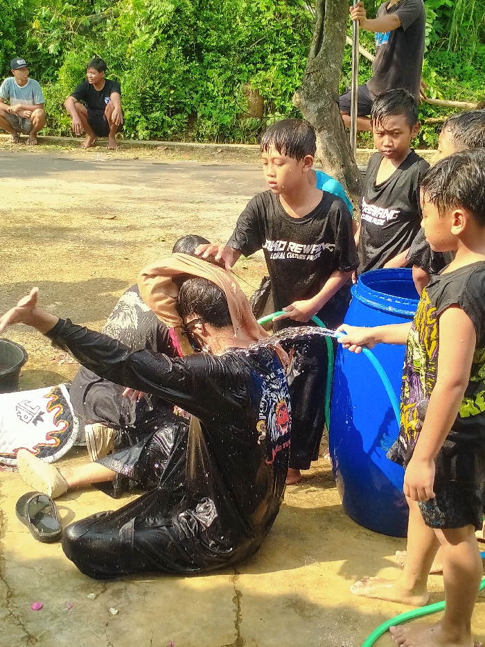 In Gebyog, Indonesia, an outdoor scene depicts a group of individuals engaged in a water activity. A male subject, wearing a black long-sleeved shirt and black pants, is seated on the ground, thoroughly saturated with water. A light brown cloth covers his head, and eyeglasses are visible. Water splashes off his back as a stream of water from a green garden hose, held by a young male in a black t-shirt, impacts him. Another young male in a black t-shirt stands near the doused subject's head. A third young male, barefoot, holds the green hose connected to a tall blue plastic barrel, which functions as a water source. Several other young males, also wet and in black t-shirts, are present around the central action.
In the mid-ground, two males are squatting barefoot on a dirt path, observing. One wears a light blue t-shirt and grey shorts, while the other wears a black t-shirt and black shorts. Further back, a male in a black t-shirt is seen holding a slender metal pole near a tree trunk.
The environment consists of unpaved, light brown ground with wet areas from the water activity. A narrow paved path or road is visible in the background, bordered by dense green foliage and trees. A grey plastic bucket, a pair of black sandals, and a white cloth with red and orange patterns lie on the ground. The scene is illuminated by bright daylight.