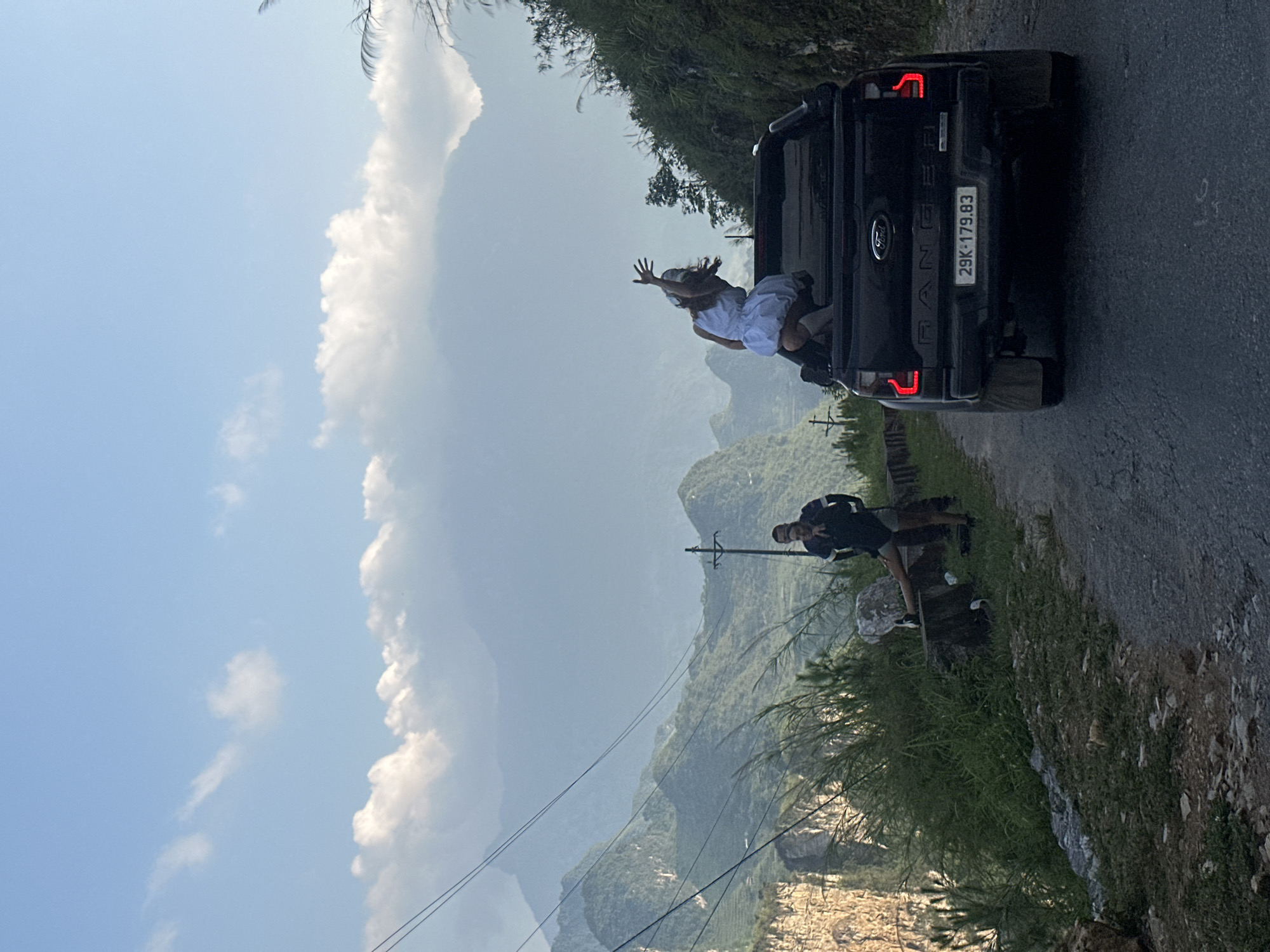 A black Ford Ranger pickup truck is parked on a road in Đường Thượng Commune, Vietnam, with the license plate 29K-179.83 visible. A woman wearing a white dress is standing in the back of the truck, waving. Another individual is standing on the side of the road taking a picture of the woman. The background consists of a mountainous landscape with a sky with clouds, and an electrical pole with wires. The road is asphalt.