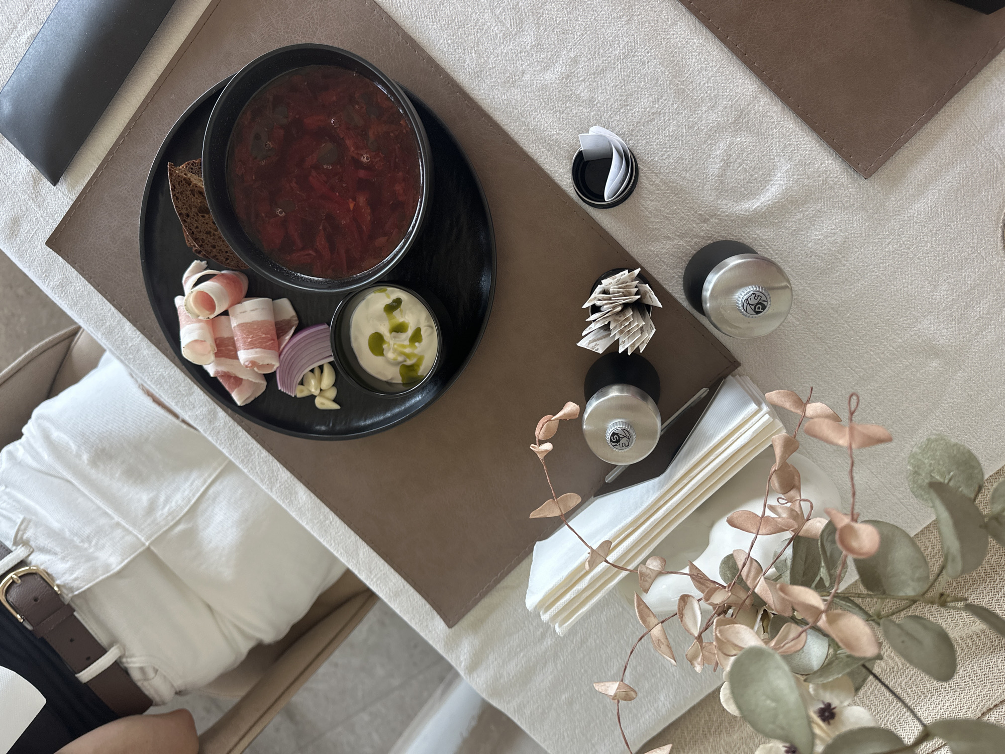 A top-down shot depicts a meal setup on a table, possibly indoors. The main focus is a black plate containing a bowl of borscht, alongside what appear to be cured meats, sliced red onion, garlic cloves, dark bread, and a small bowl of sour cream garnished with green oil. Cutlery is placed on top of a pile of white napkins. Two salt and pepper shakers are next to the napkins. Nearby are small containers of toothpicks and folded paper. The table is covered with a light-colored tablecloth. Parts of a chair, a brown leather belt, and a person's leg are partially visible in the lower left corner of the frame. A floral arrangement sits on the edge of the table. The photograph was likely taken in Boreti, Montenegro.