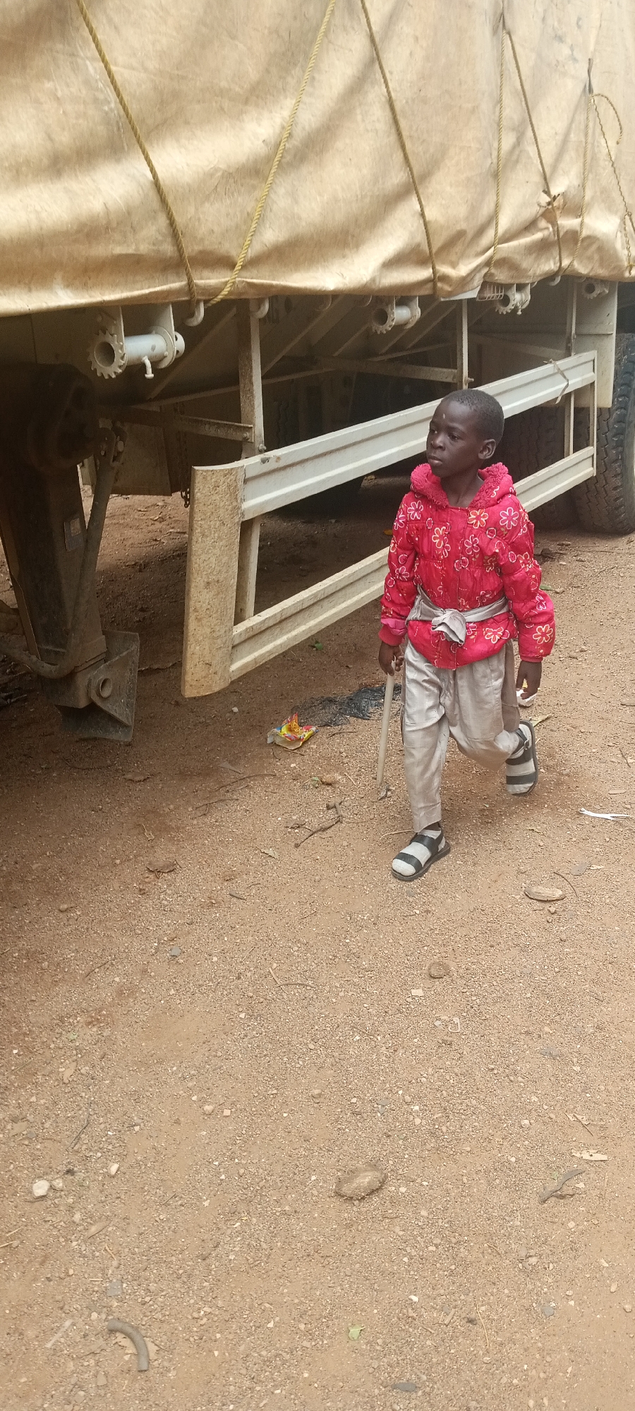 A young person stands in a dirt-covered area next to a truck. The individual wears a red floral-patterned jacket, light-colored pants, and striped sandals, and carries a stick. The truck has a light-colored tarp secured with ropes, and its undercarriage is visible. The scene is filled with dirt and some debris.