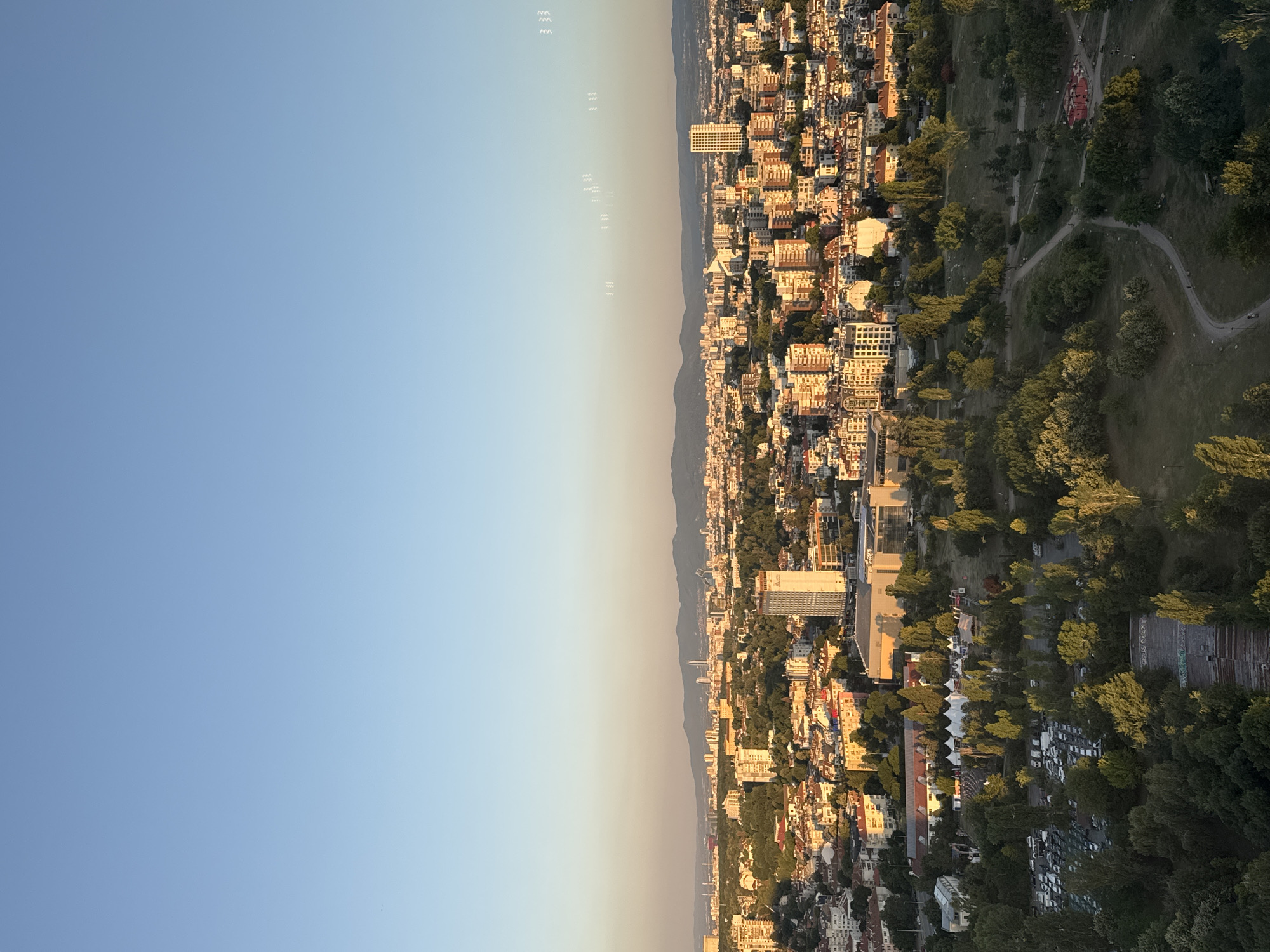 This is an aerial, high-angle, wide shot of Sofia, Bulgaria, taken during golden hour. The city is spread out below with a clear, light blue sky dominating the upper portion of the image. The sun is low on the horizon, casting long shadows and illuminating the buildings with a warm, orange glow. In the foreground, a green park area with winding paths and scattered trees leads into the urban landscape. The mid-ground features a dense collection of residential and commercial buildings, many with red-tiled roofs. Several taller buildings, including what appears to be a hotel or office building with a prominent clock face, punctuate the skyline. In the distance, a range of mountains forms a hazy backdrop. There are no visible people or specific actions taking place. A parking lot filled with vehicles is visible in the lower left portion of the image, adjacent to the park. The overall impression is a peaceful urban panorama at dusk.