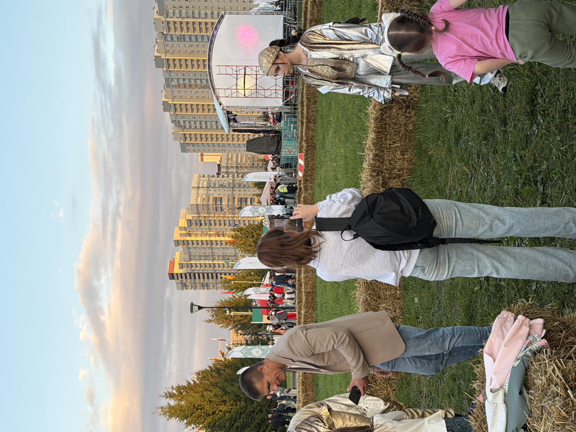 An outdoor festival scene unfolds at dusk, with a stage in the background and high-rise buildings beyond. In the foreground, a man in a beige blazer and jeans holds a phone, looking down. To his right, a woman with a backpack is taking a picture of a performer dressed in a shimmering silver outfit. A young girl in a pink t-shirt and green pants stands facing away from the camera, looking towards the performer. Hay bales are arranged in the grassy area, with some clothing items folded on one of the bales. Flags and tents are visible in the mid-ground, suggesting a public event. The sky is partly cloudy with the soft hues of sunset.