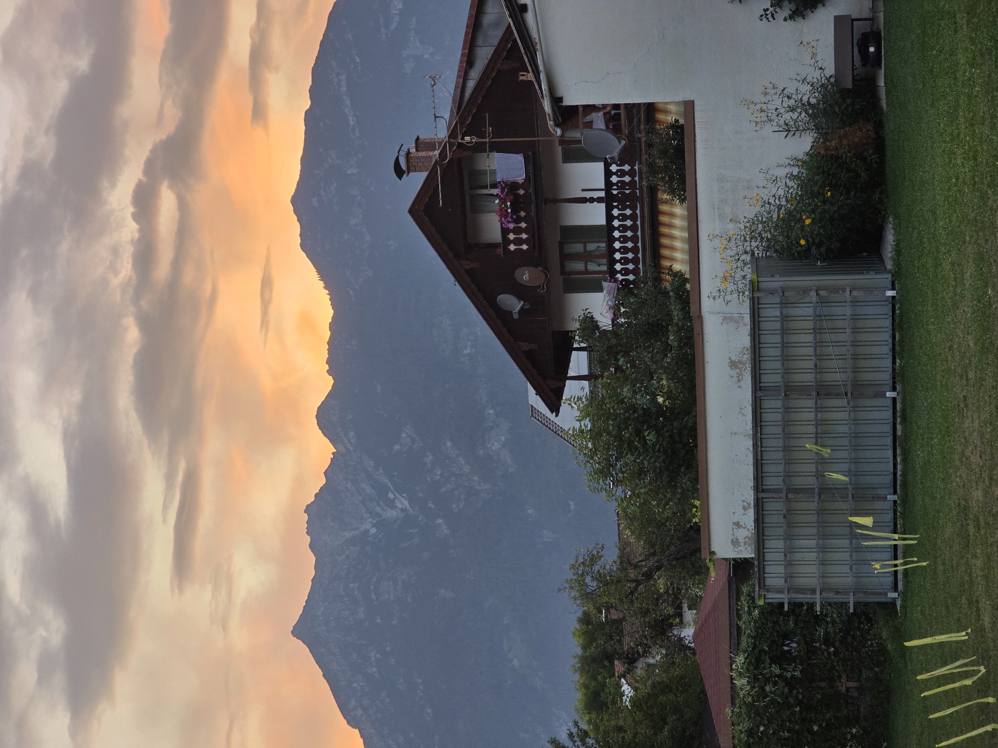 The image depicts a picturesque sunset scene in Garmisch-Partenkirchen, Germany, with the majestic Alps serving as a dramatic backdrop. In the foreground, a traditional Bavarian house, characterized by its dark wood trim and intricate balcony railings, stands bathed in the warm, golden light of the setting sun. The sky is a canvas of soft oranges, pinks, and grays, with clouds casting a gentle glow over the mountainous landscape.
The house features a steeply pitched roof, a prominent chimney, and a balcony adorned with colorful flowers. Several satellite dishes are visible on the exterior, suggesting modern amenities within the historic architecture. A white wall separates the property from the street, and a neatly manicured lawn stretches out in front of it.
In the middle ground, a greenhouse-like structure with a metal frame and translucent panels is situated, with yellow ribbons or markers tied to a wire running across its front. Adjacent to it, a dense cluster of green bushes and wildflowers, including bright yellow blossoms, adds a touch of natural beauty. A dark-colored park bench is positioned to the right of the structure, with a black object, possibly a trash receptacle, beneath it.
The background is dominated by the imposing silhouette of the Alps, their rugged peaks softened by the atmospheric haze of twilight. The scale of the mountains, contrasted with the charming human dwelling, creates a sense of both grandeur and tranquility. The overall atmosphere is one of serene beauty, capturing the essence of a peaceful evening in the Bavarian Alps.