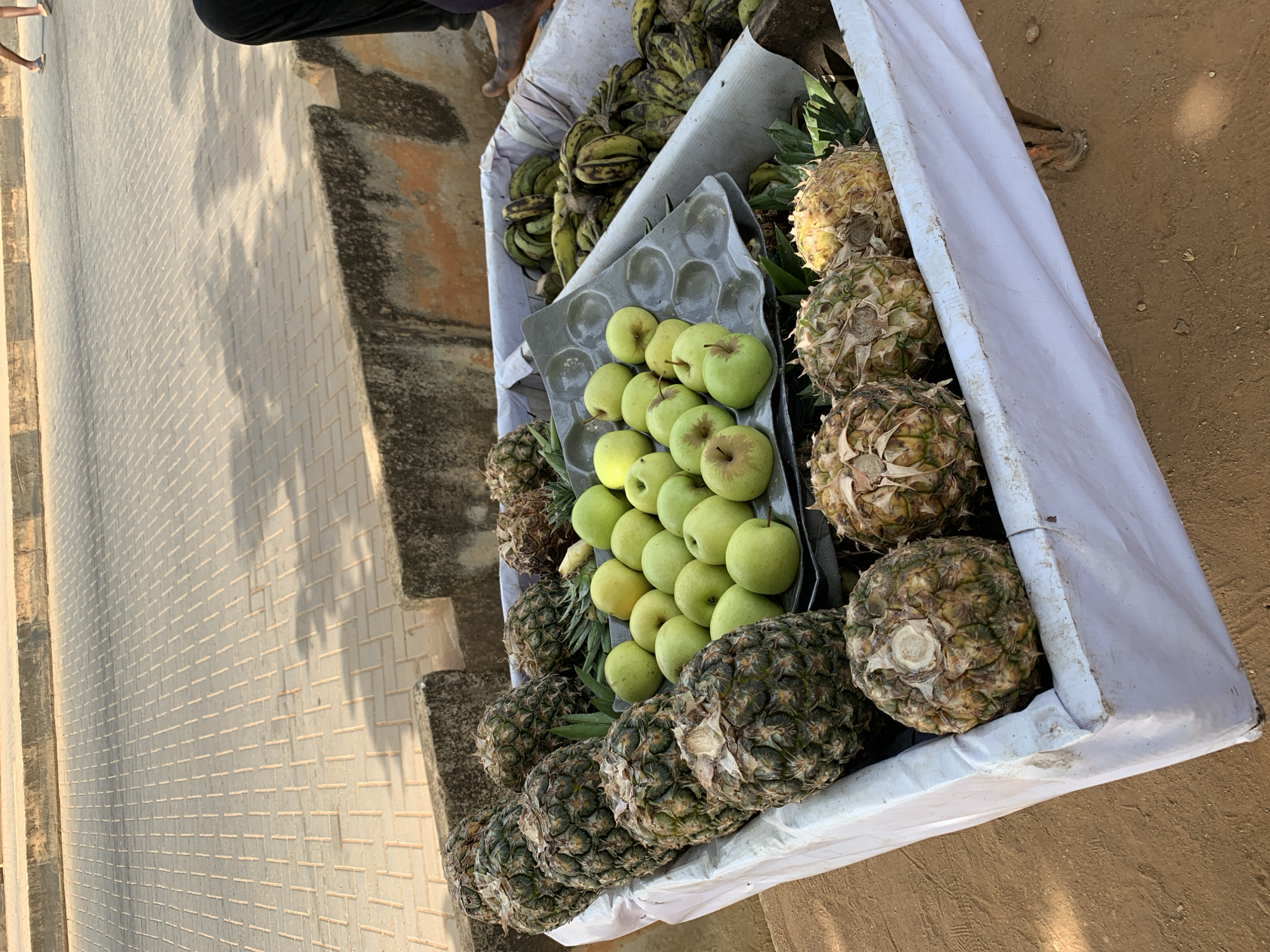 A high-angle, medium shot shows a white rectangular vendor's cart filled with produce: pineapples, bananas, and green apples. The pineapples, in the foreground and right side, display varying shades of green and brown. The green apples are centrally positioned in a gray molded plastic tray, sitting amongst the pineapples. Bunches of green and yellow bananas are situated towards the back of the cart. The cart rests on the right edge of a brick-paved area adjacent to a sandy surface. Part of a person's legs and feet are visible at the top left corner of the frame. The scene is located in the city of Ikorodu, Nigeria.