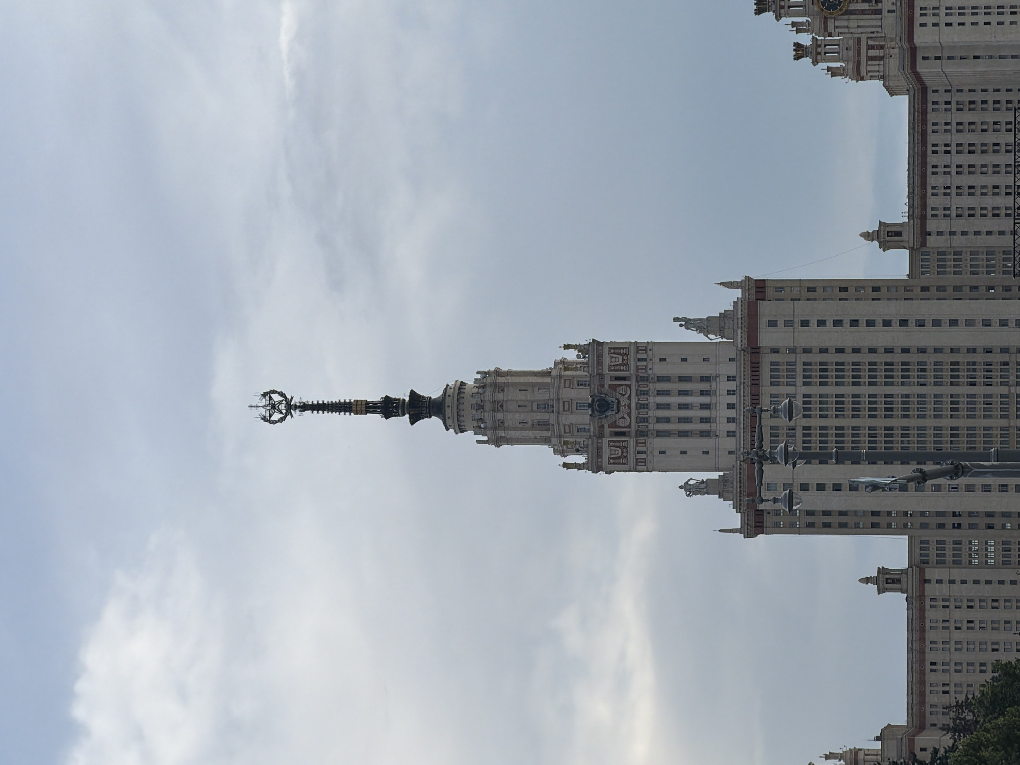 This is a vertical, daytime, outdoor photograph of a large, ornate building. The building is tall and made of light-colored stone, with many windows and decorative elements. The style of architecture appears to be neo-Gothic or Stalinist, with spires and ornate detailing. The building is partially obscured by a light gray sky with some wispy clouds.
The primary subject is the building itself, which dominates the frame. It appears to be a grand structure, possibly a university building or a government edifice. There are no people visible in the photograph.
Notable details include the intricate spires and the many repetitive windows, suggesting a large capacity or a monumental purpose for the building. A prominent spire with a star-like ornament at its apex is visible towards the top of the frame, in the center-left. The lighting suggests it is daytime, possibly late morning or early afternoon, with diffused light indicating a partly cloudy or overcast sky.
There is no visible text in the image. The scene conveys a sense of grandeur and architectural impressiveness. The orientation of the image is vertical, and the perspective is from ground level looking up.