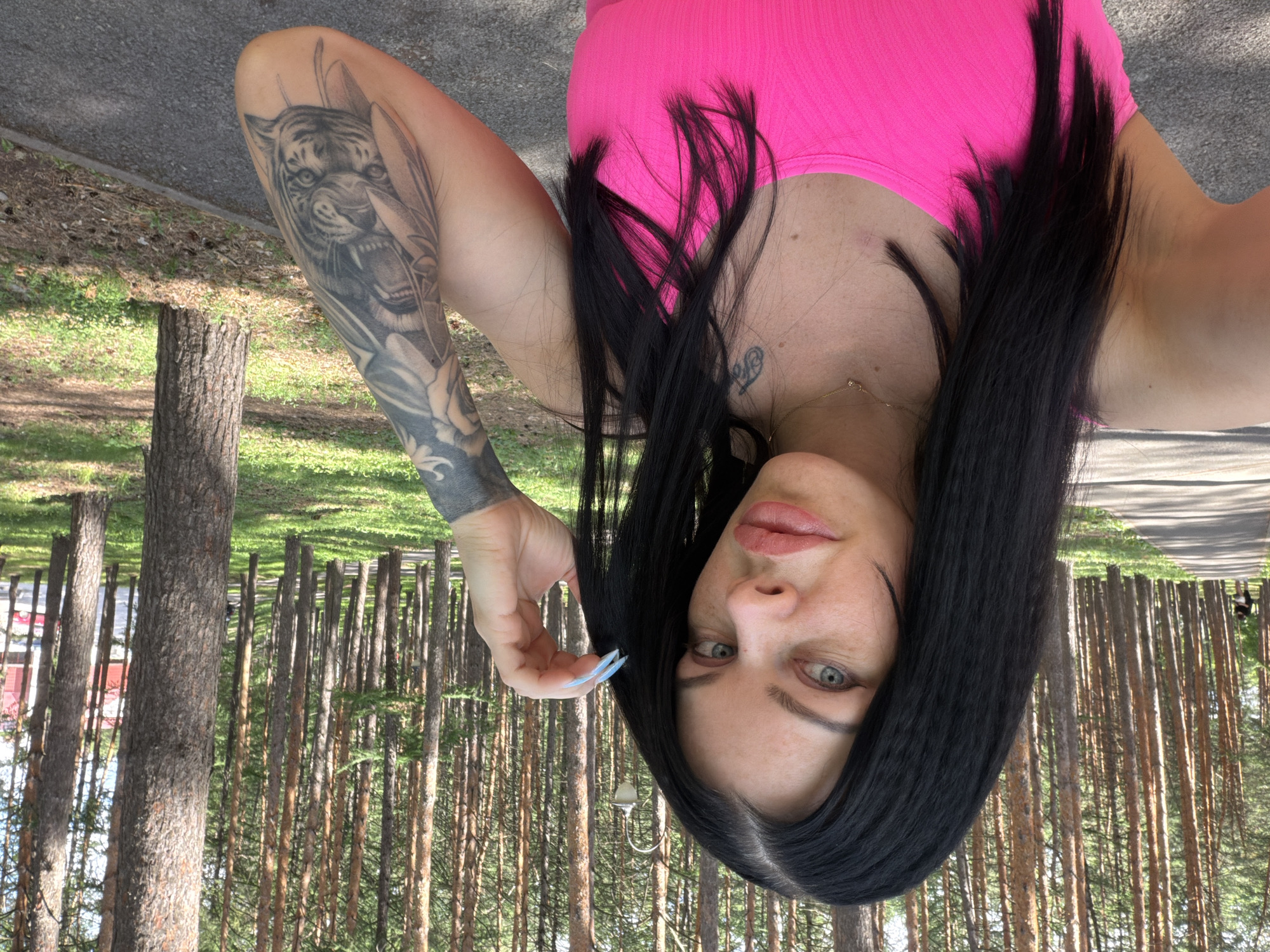 The media file shows a person lying on their back outdoors. They are wearing a bright pink top. The person has long, dark hair and a tattoo of a tiger on their left arm. Their face is turned towards the camera, and they have light-colored eyes. The background consists of a forest with many tall, thin pine trees and green grass. There is also a paved path visible. The lighting suggests it is daytime, and the overall atmosphere seems relaxed and casual. There is no visible text in the image. The scene appears to be in Ufa, Russia.