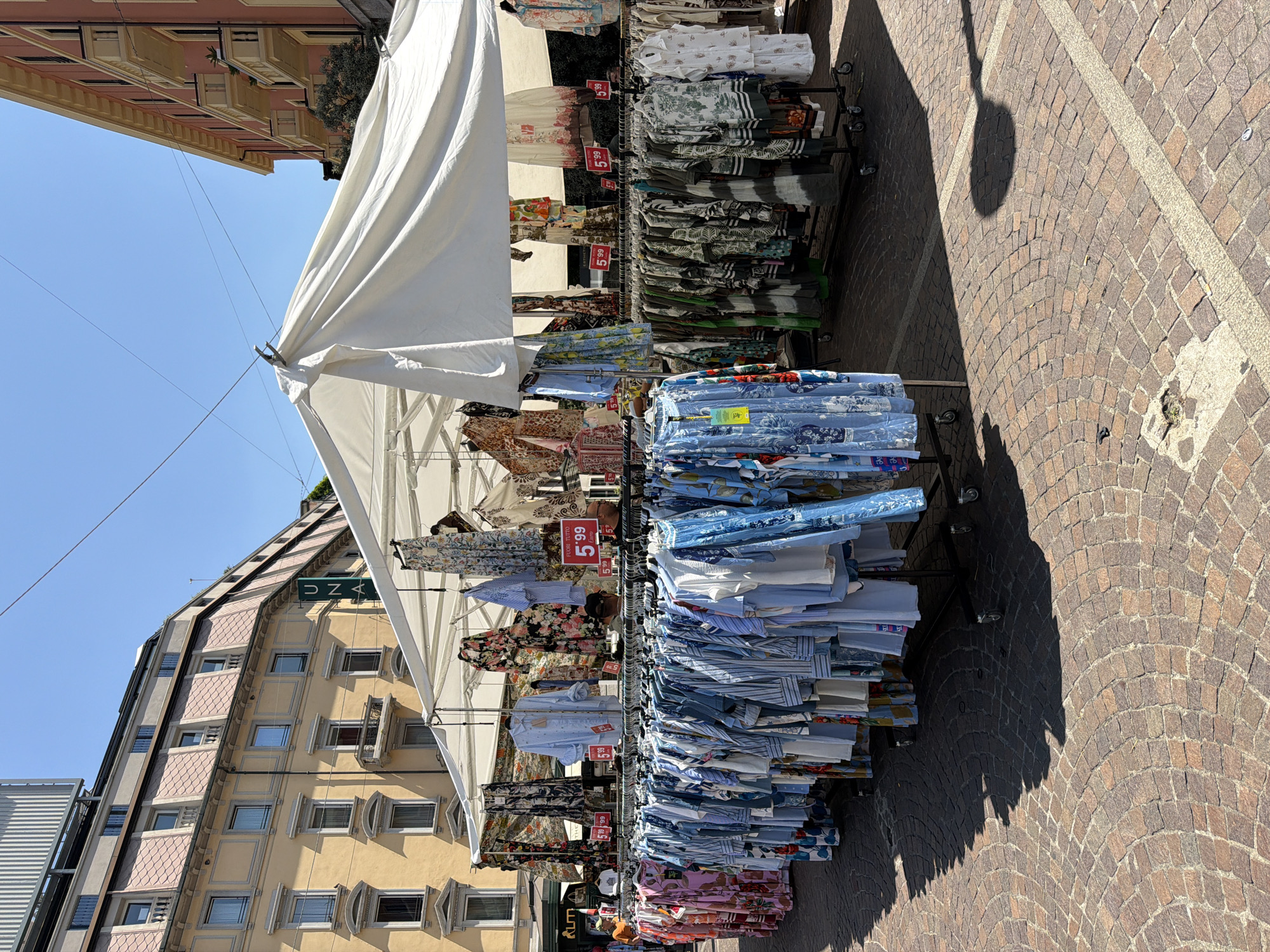 An outdoor clothing market is set up under a white canopy in Milan, Italy. The market displays a variety of women's clothing, primarily dresses and blouses, organized on racks. Some garments feature floral patterns, while others are striped or solid blue. Several price tags are visible, indicating prices of "5,99 Euro" and "5,99". In the background, buildings with yellow and pink facades line a street paved with cobblestones. The sky is clear and blue, indicating a sunny day.