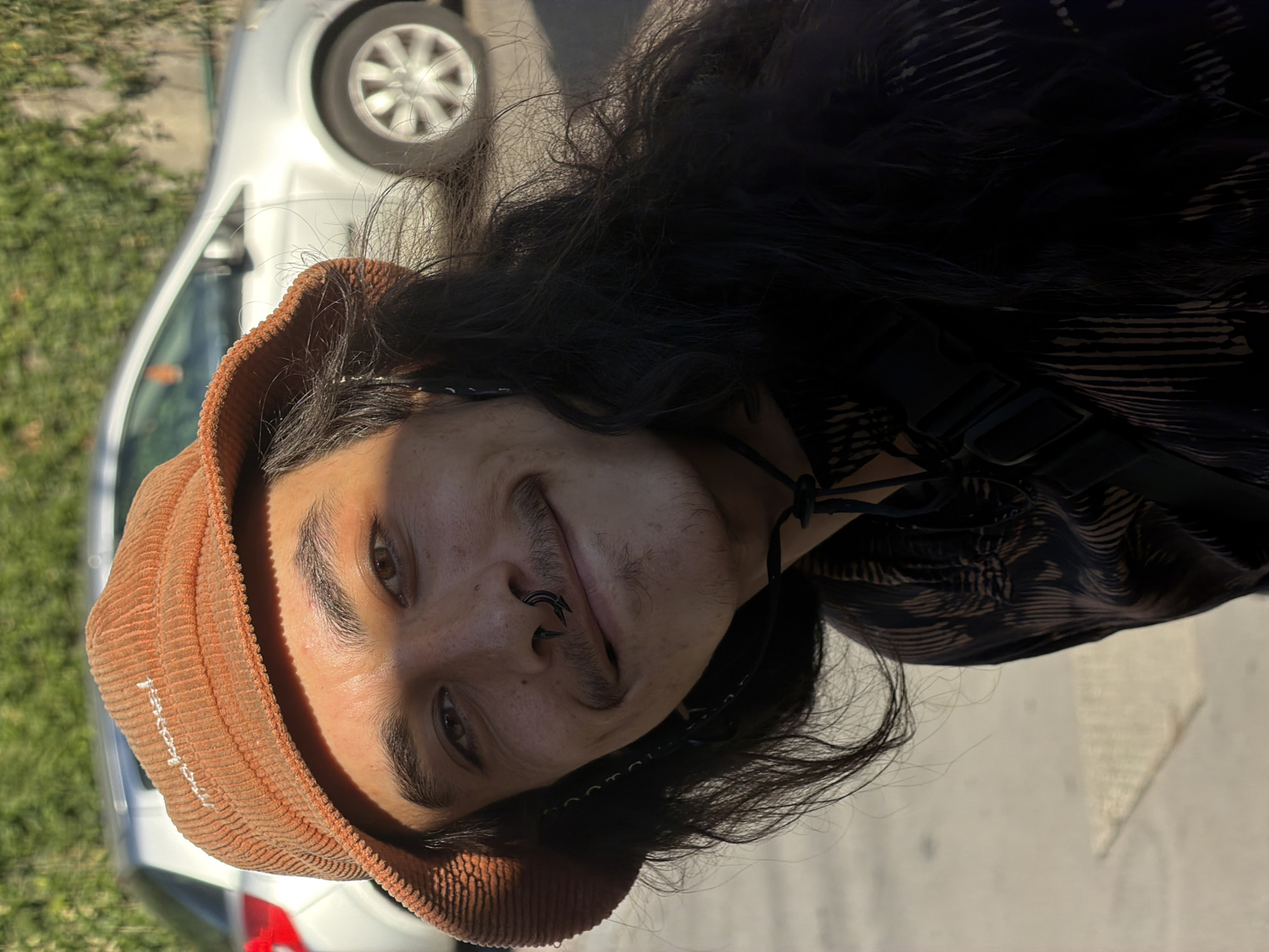 A close-up, head-and-shoulders shot captures a young person with long, dark, wavy hair, wearing an orange corduroy bucket hat and a dark patterned shirt. They have a septum piercing and a thin mustache and goatee. The person is looking directly at the camera with a slight smile. In the background, a silver car is parked on a street, with a blurred green hedge and a building facade visible. The lighting suggests it is daytime, with sunlight casting shadows on the person's face and hat. There is no indication of the location being Lyon, France.