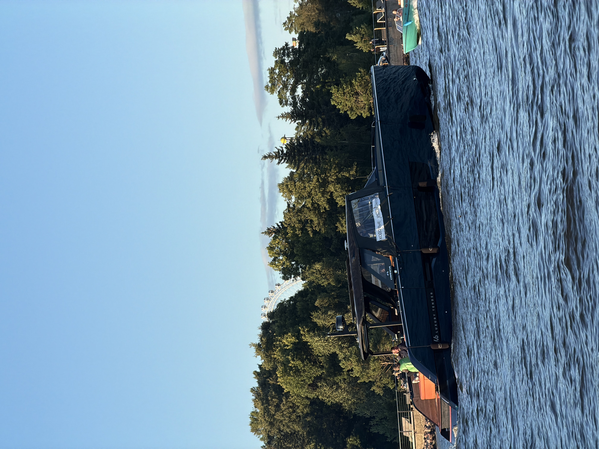 The image captures a scene on the Neva River in Saint Petersburg, Russia, during daylight. A large, dark-colored luxury tender is the primary subject, moving through the water, creating ripples. The boat has a covered cabin area with several people visible inside and on the back deck. The name "LUXURY TENDER" and the number "46" are visible on the hull. To the right of the tender, a smaller, green boat with two people is also in motion. In the background, a dense line of green trees borders the riverbank, with a Ferris wheel partially visible above the treeline, suggesting the presence of an amusement park or recreational area. The sky is a clear, light blue.