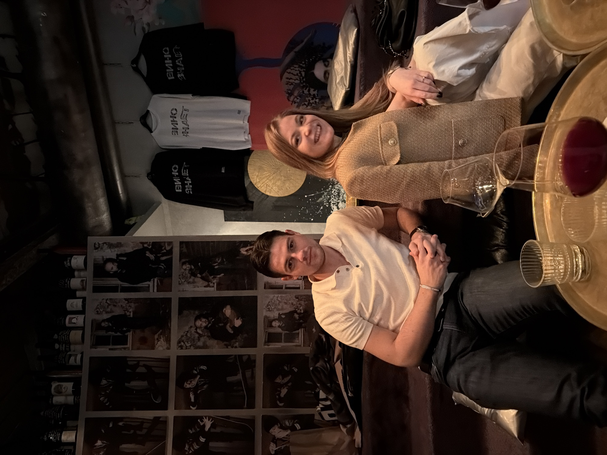 The image depicts a man and a woman seated indoors, likely in a bar or lounge in Moscow, Russia, suggested by the "ВИНО ЗНАЕТ" (Wine Knows) graphic visible on apparel hanging on the wall. The woman has long blonde hair, wears a tan tweed jacket, and is smiling. The man wears a light-colored polo shirt and dark jeans, and is also smiling. They are positioned close to each other, possibly on a couch or lounge seating. A small gold-colored table in the foreground holds a wine glass filled with red wine, another empty wine glass, and two textured drinking glasses. Behind them, on the wall, are three hanging shirts with "ВИНО ЗНАЕТ" printed on them and a series of framed monochrome photographs. A wine rack stacked with wine bottles is also visible. There is an abstract painted mural on the wall behind the seating area.