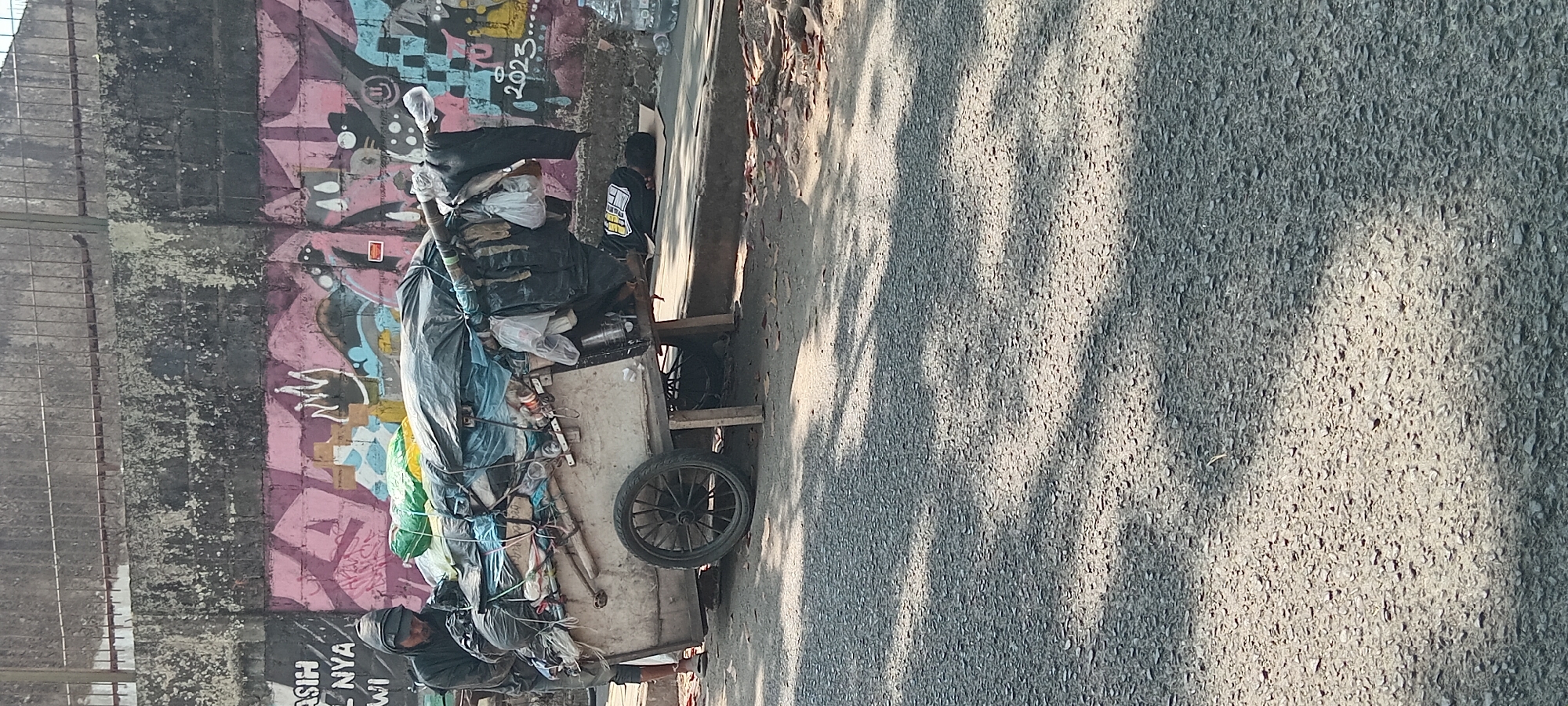 The image depicts a street scene in Tasikmalaya, Indonesia. A person wearing a dark hooded jacket and pants is seen pushing a cart laden with various items, including plastic bags, tarps, and possibly recyclables. The cart has a single wheel and appears to be made of wood and metal. Behind the cart, a graffiti-covered wall forms the backdrop, with artistic designs and what appears to be the year "2023" painted on it. The ground is paved with asphalt, and shadows suggest the presence of overhead structures or trees. Another person is lying down on a piece of cardboard on the sidewalk next to the cart, appearing to be asleep. The overall scene suggests an urban environment where street vendors or individuals collecting discarded materials operate.