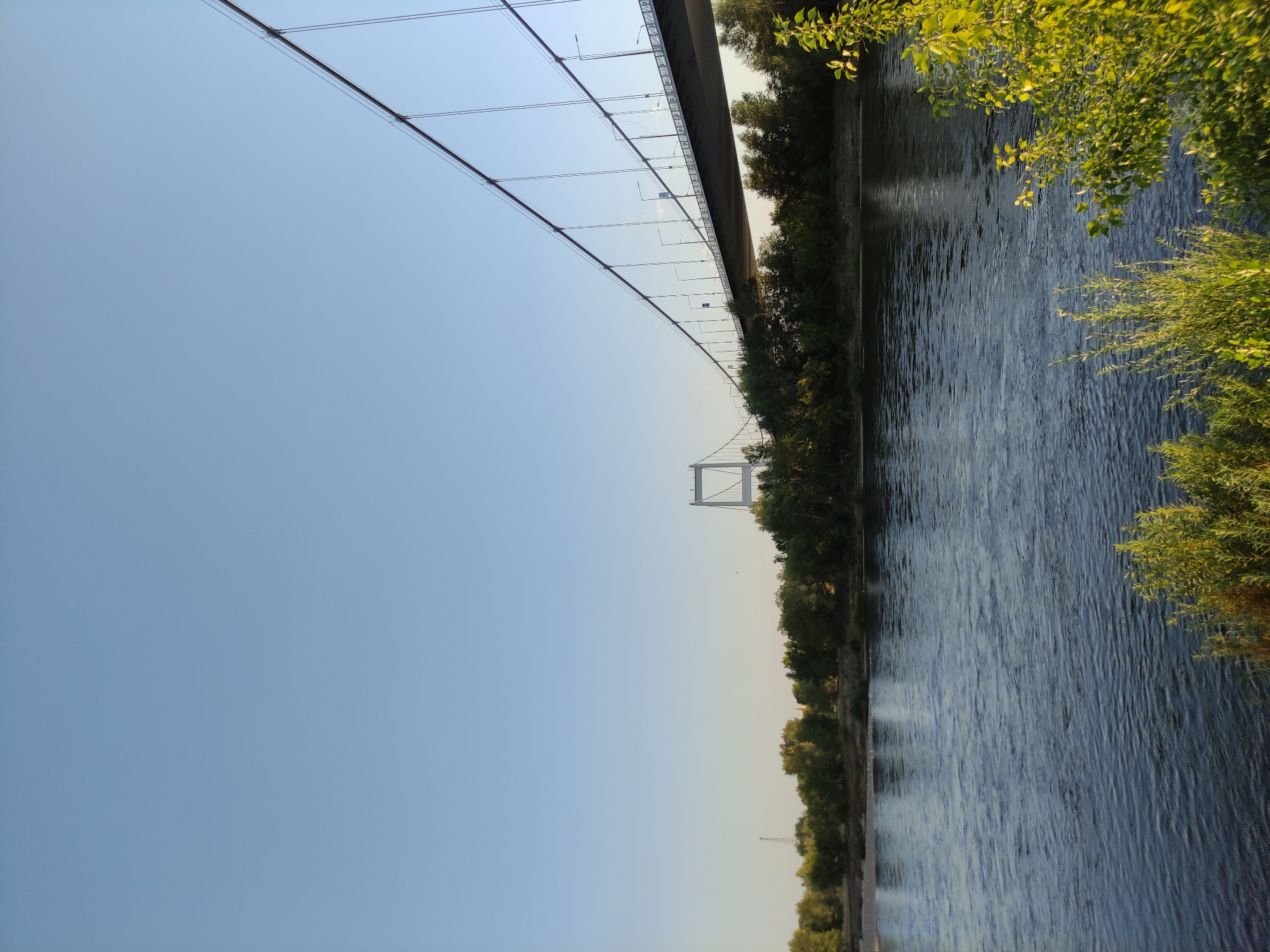 The image shows a wide river with a suspension bridge crossing it. The sky is clear and blue, and the sun appears to be low on the horizon, casting a warm glow on the scene. On the far bank of the river, there is a line of trees, and further in the distance, a tall radio tower can be seen. The bridge itself is a prominent feature, with its sleek design and numerous suspension cables stretching across the frame. In the foreground, the water of the river is visible, with gentle ripples on its surface. The overall atmosphere is serene and picturesque, with a sense of open space and natural beauty. This scene likely depicts the Irtysh River and the suspension bridge in the city of Semey, Kazakhstan.