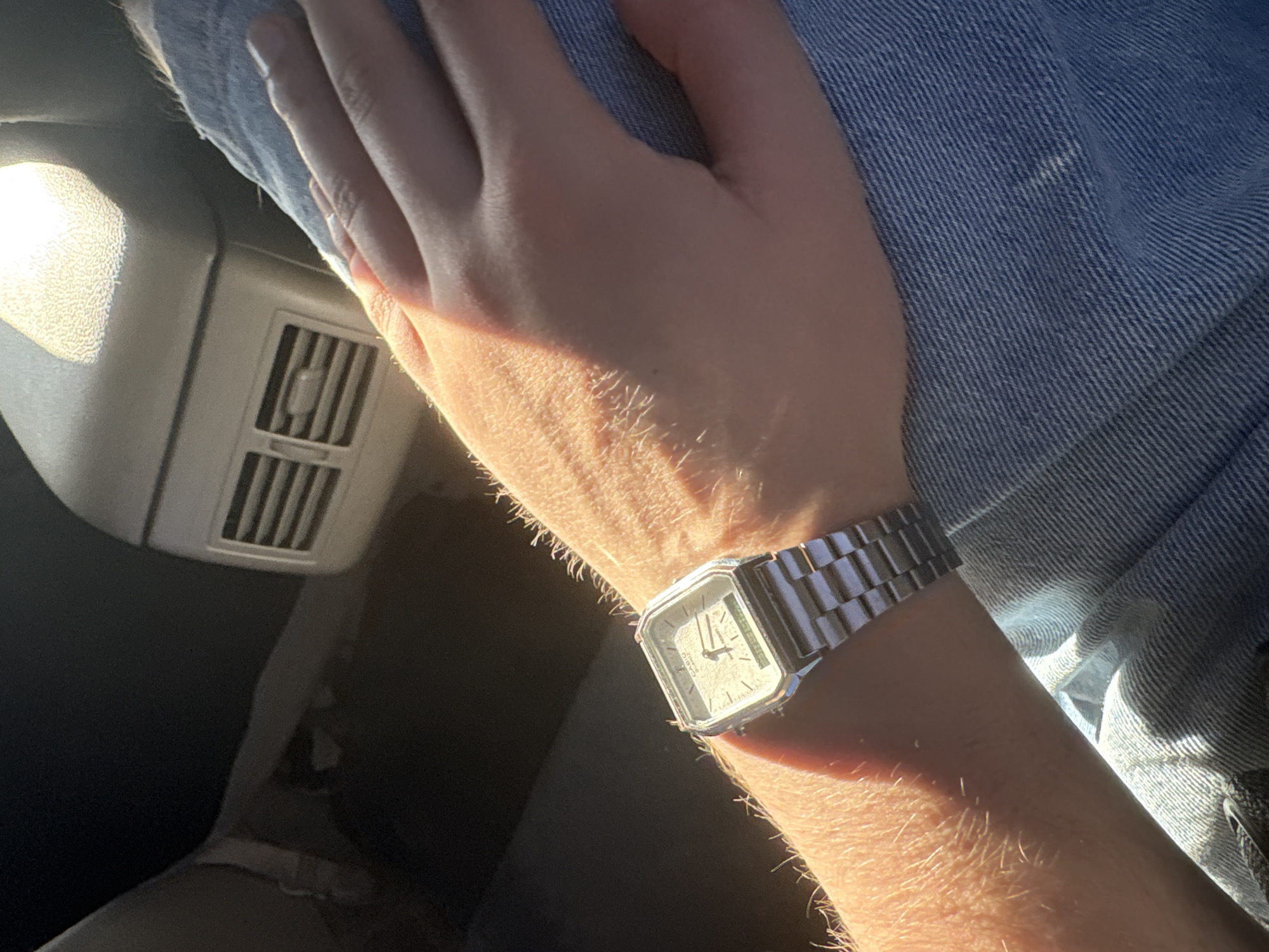 The image shows a close-up of a person's left arm and wrist, adorned with a silver digital watch with a square face. The arm is hairy and bathed in warm, directional sunlight, casting shadows and highlighting textures. The person is wearing blue denim jeans. In the background, blurred, is the interior of a vehicle, possibly a car, with a vent on the back of a seat visible, suggesting a seated position inside a car. There is no identifiable indication of the city of Dnipro, Ukraine in the image.