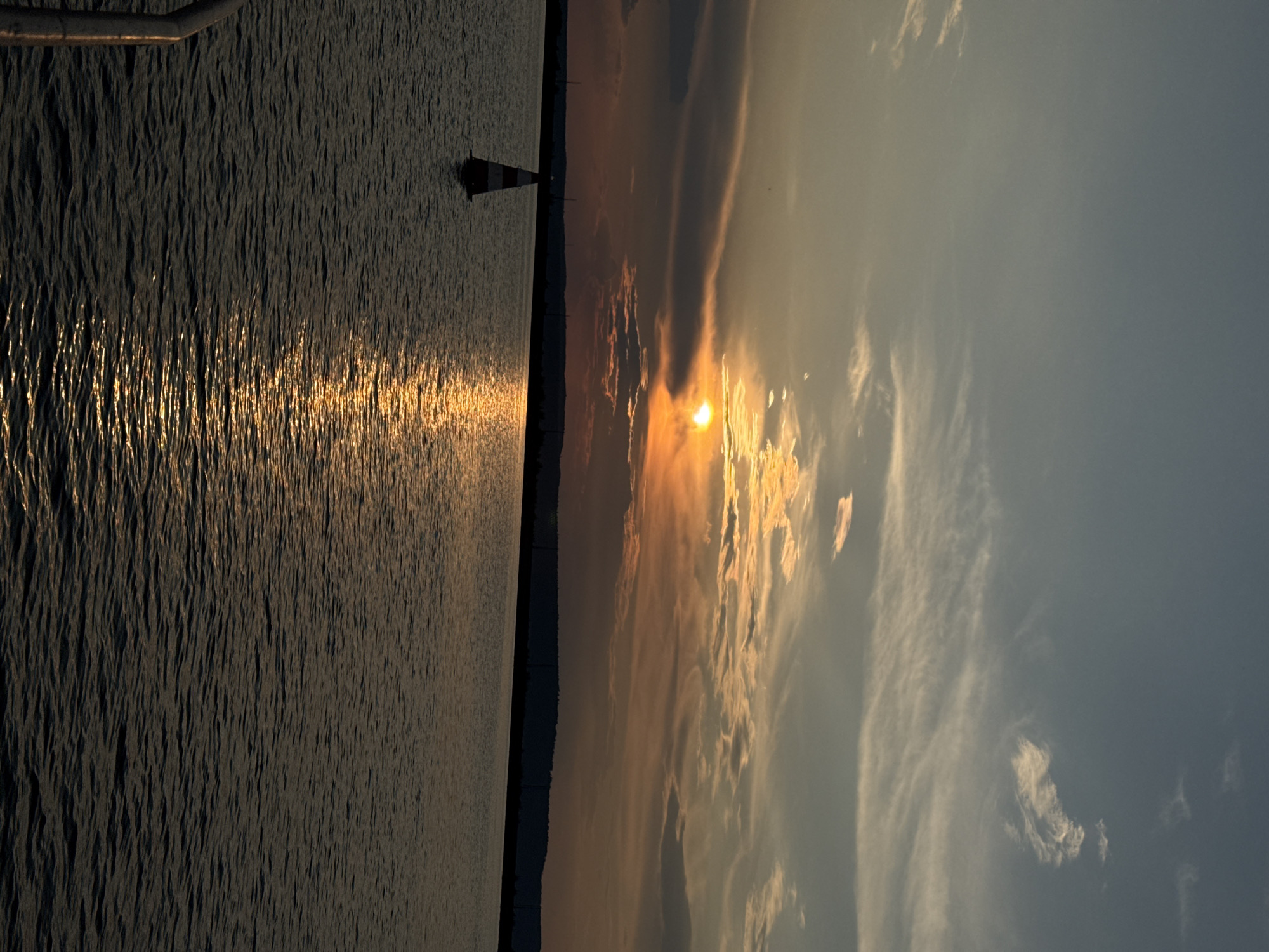 The image shows a sunset over the Kama River in Chaikovsky, Russia. The sun is a bright orange orb low on the horizon, partially obscured by clouds. The sky is a gradient of blue, orange, and yellow, with wispy clouds stretching across it. The water of the river is dark and rippled, reflecting the colors of the sky. In the foreground, a buoy with red and white stripes stands in the water. In the background, the silhouette of a distant treeline is visible against the horizon. A metal railing, likely from a boat or pier, is visible on the left side of the frame. The scene is calm and serene, with the natural beauty of the sunset dominating the image.