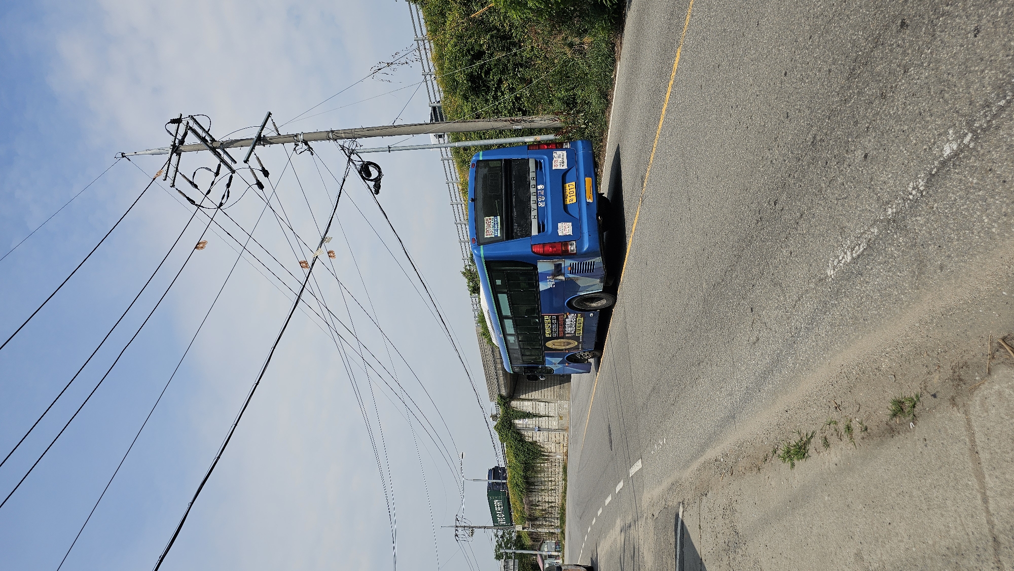 A blue Hyundai bus with Korean text and a license plate reading "경기 1017" is driving on a two-lane asphalt road with a yellow dividing line. The bus is in the right lane. A concrete retaining wall covered in ivy is to the left of the road, with an overpass in the background. There are electrical poles and overhead wires crossing the scene. The sky is blue with some clouds. In the distance, a green truck is visible. The image appears to be taken in Pyeongtaek-si, South Korea.