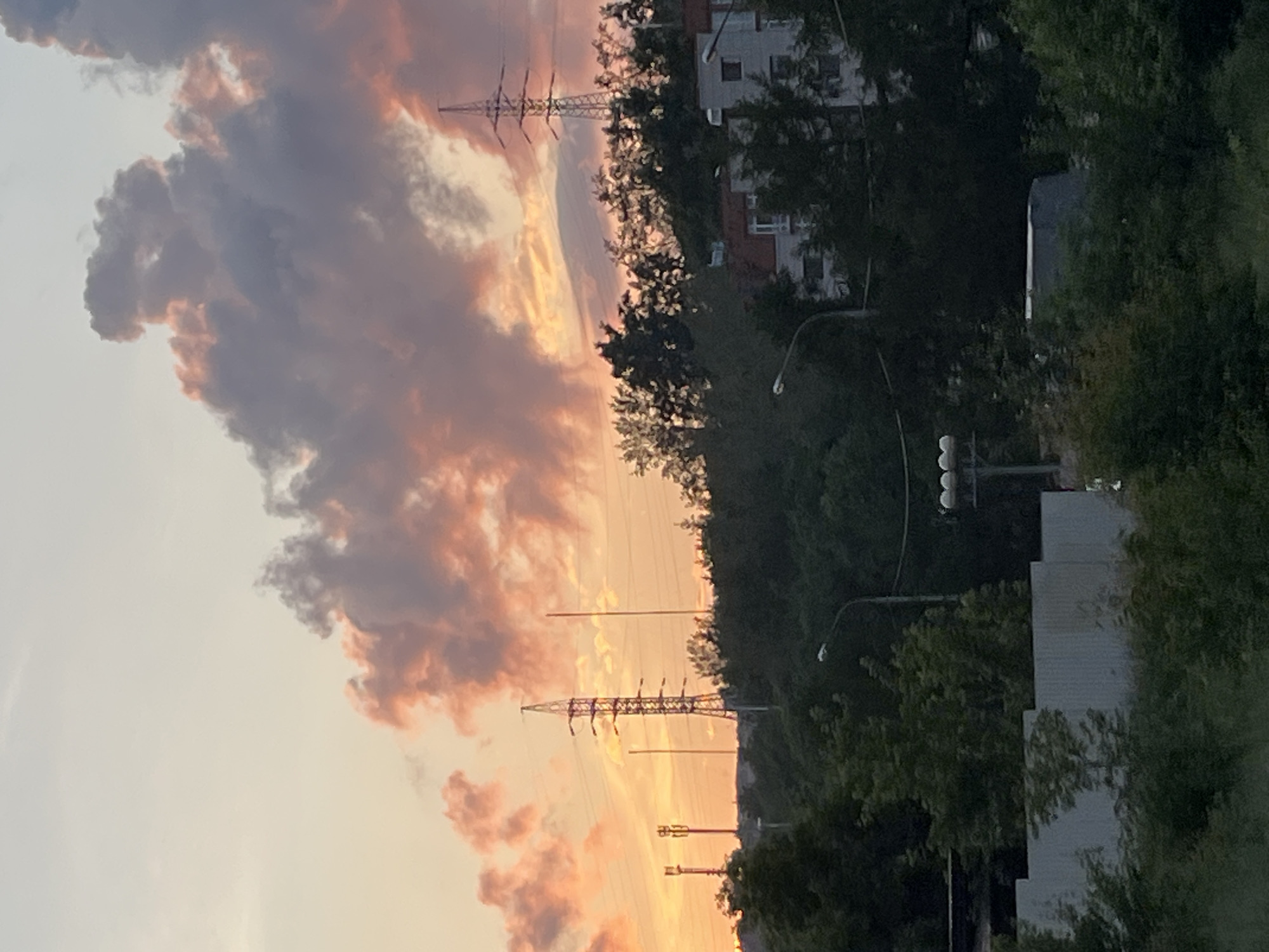 The image depicts a twilight scene in Krasnoyarsk, Russia. The sky is a gradient of pale yellow and orange, with large, textured clouds tinged with pink and purple hues, suggesting the last light of sunset. Dominating the middle ground are a series of silhouetted green trees on a hill, with buildings partially visible through the foliage. Several power lines and transmission towers are prominent in the upper portion of the image, indicating an urban or semi-urban environment. In the foreground, a corrugated metal fence runs horizontally across the lower part of the frame, partially obscured by greenery. The overall atmosphere is one of a calm, overcast evening.