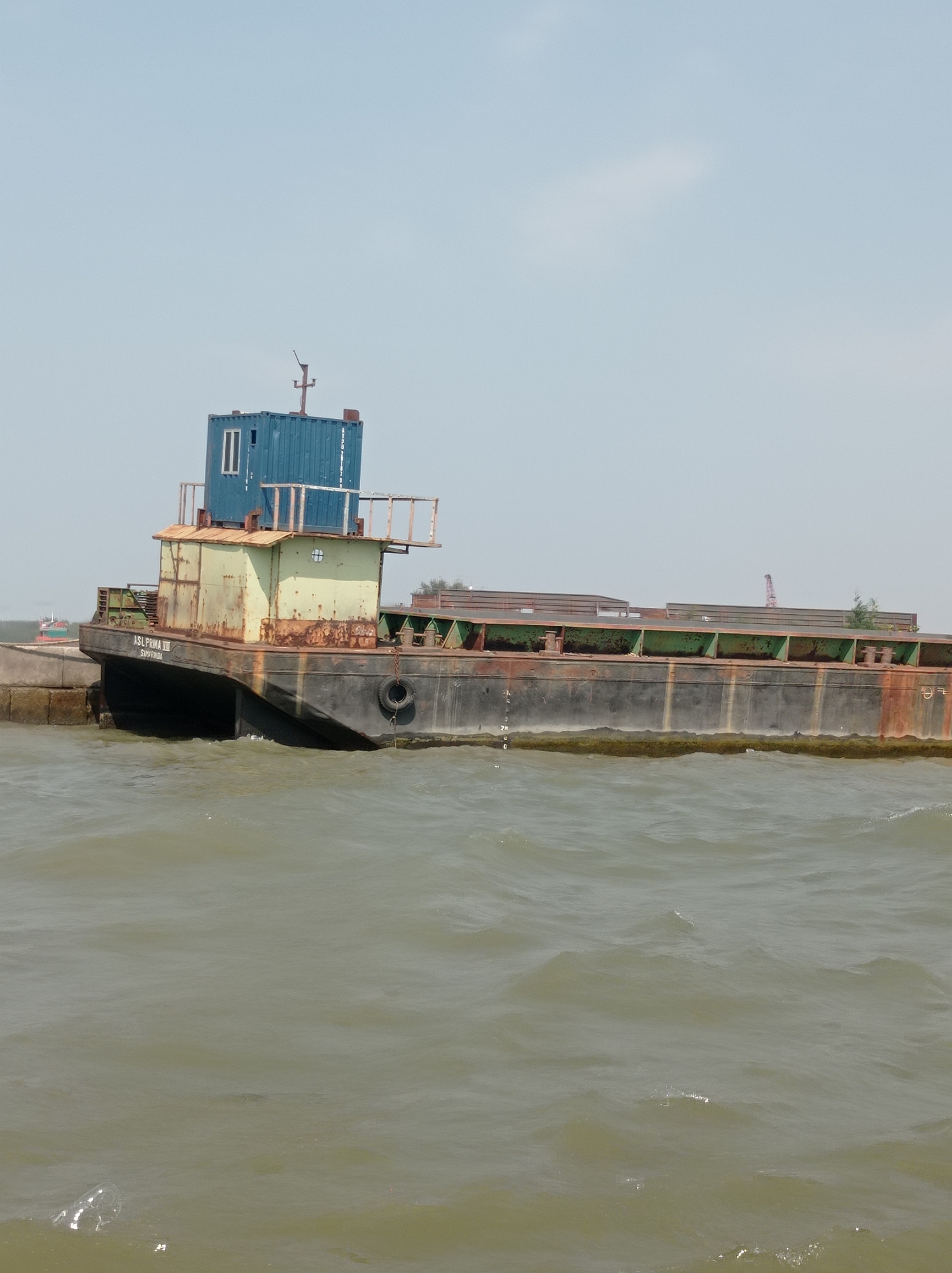 A side view of a rusty, utilitarian barge docked at the edge of murky green water under a clear, hazy sky. The barge, identified with the name "ASL PRIMA VIII SIMAMURIDA" printed on its hull, has a weathered, metallic exterior, primarily black with green and rust-colored accents. On its deck, a blue shipping container is stacked on top of a smaller, pale green structure. The structure on the barge has visible windows and railings. The water around the barge is choppy, with small waves creating ripples. The background shows a distant, hazy coastline with hints of vegetation and industrial structures, including what appears to be a crane. The scene suggests a working port or industrial waterway, likely in Tegal, Indonesia, given the presence of the named barge.