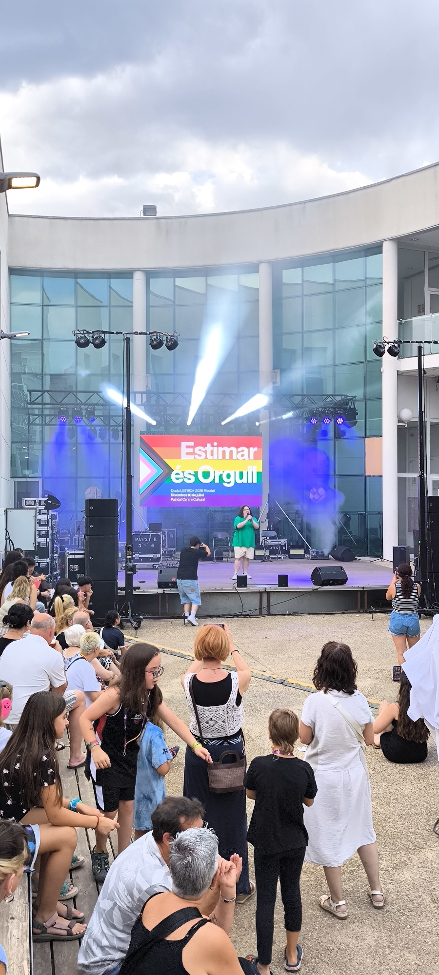 The image depicts an outdoor event with a stage set up against a building with large glass windows. On the stage, a screen displays the text "Estimar és Orgull" in colorful letters, along with smaller text indicating "Dia Lesbi+ 2026 Ripollet Divendres 10 de juliol Pati del Centre Cultural." Two performers are on stage, one in a green top and shorts speaking into a microphone, and another person in a black t-shirt and denim shorts standing to the left. The stage is lit with blue and white lights, creating a hazy atmosphere. In front of the stage, an audience of various ages is seated on wooden bleachers and standing on the ground, many of whom are looking towards the stage. Several audience members are holding up phones, seemingly to take pictures or videos. The environment suggests a public gathering or festival held at the Pati del Centre Cultural in Ripollet, Spain, on July 10th, 2026, celebrating LGBTQ+ pride.