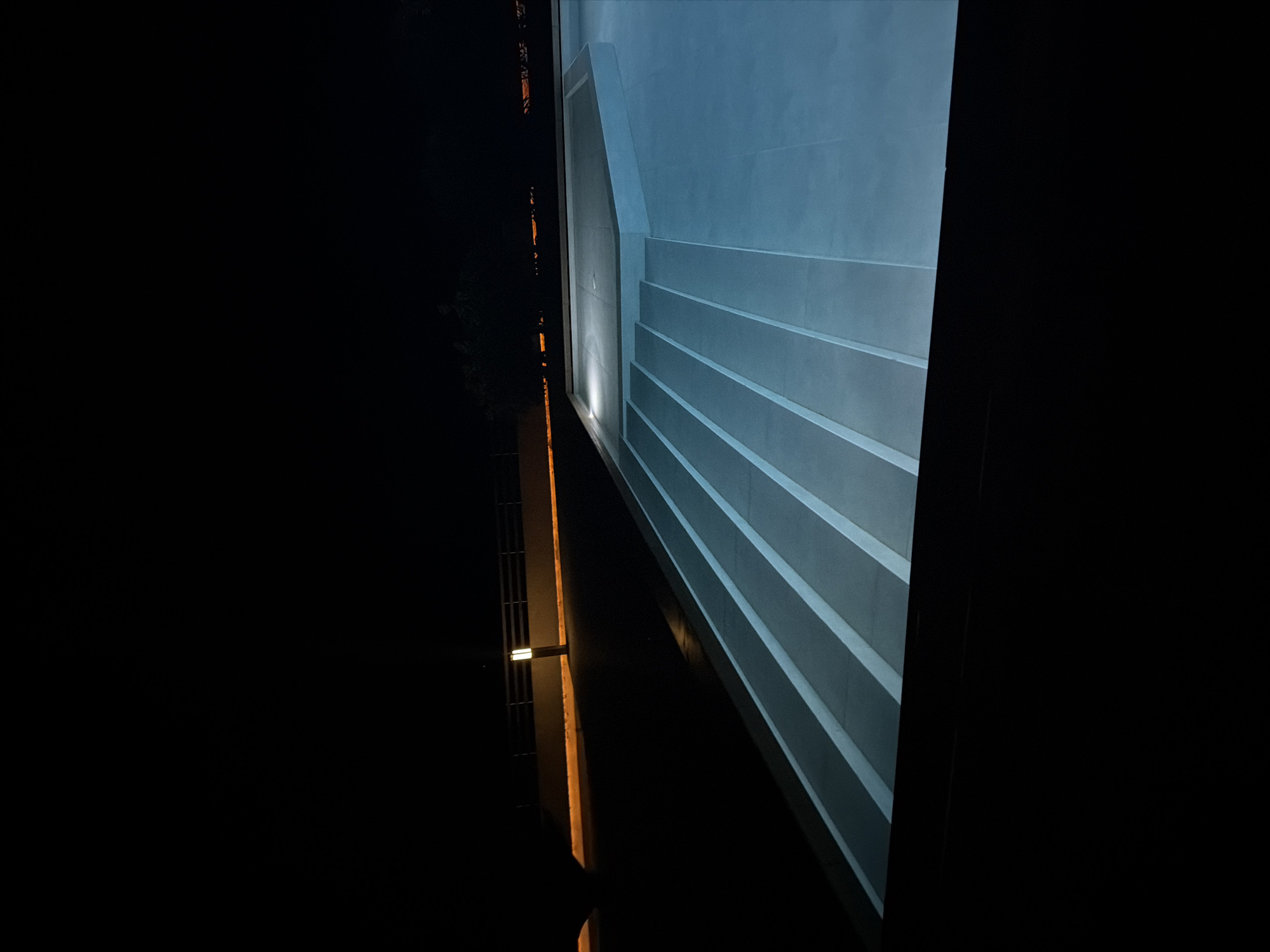 A wide shot captures an outdoor swimming pool at night, illuminated by soft, blue underwater lights. The pool features a tiered, illuminated staircase on the left side, leading down into the water. The surrounding area is dark, with a fence and a single post lamp visible in the background. The scene is tranquil and serene, suggesting a private or resort setting. The location is identified as Viglakia, Greece.