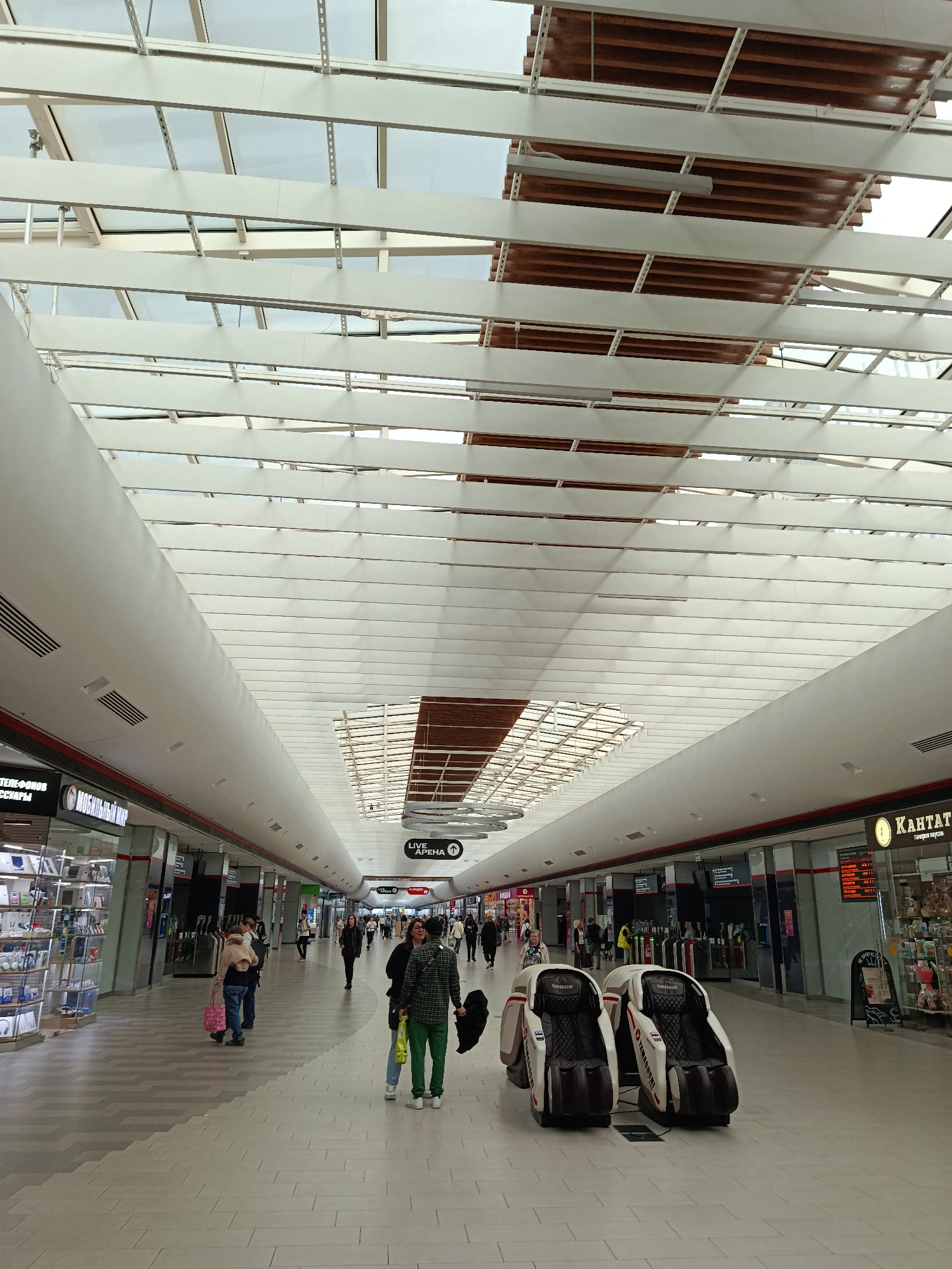 The image shows an indoor shopping mall with a high, sloped ceiling made of glass and white beams, allowing natural light to filter in. The mall features shops on either side of a wide walkway. The floor is tiled with a patterned design.
Several people are visible walking through the mall. In the foreground, two people are standing and looking at something off-camera, while two massage chairs are placed in the walkway. Further down the mall, more individuals are seen browsing shops or walking.
Shop signs are visible along the walls. On the left, a sign reads "МОБИЛЬНЫЙ МИР" (Mobile World). On the right, a sign says "КАНТАТА" (Kantata), possibly a store name. Another sign in the distance indicates "LIVE АРЕНА" (Live Arena).
The scene appears to be during the daytime, as indicated by the natural light. The overall atmosphere seems calm and ordinary, typical of a shopping center. There are no obvious indicators of the specific time of day beyond it being open for business. The weather outside is not discernible. The setting is clearly a modern retail environment.