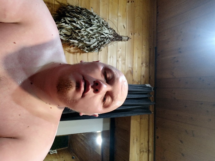 The image displays an interior scene in Nizhneye Zadolgoye, Russia, depicting a single adult male in what appears to be a sauna or banya. The individual, seen from the chest up, has a bald head and a short, reddish-brown goatee. His skin is noticeably flushed and beaded with sweat across his face, neck, and bare upper torso. His eyes are closed or heavily lidded, and his mouth is slightly ajar. The background consists of horizontally oriented light-toned wooden plank walls and ceiling. To the left of the subject, a dried venik (bath broom), made of pale green and brown leaves, is hanging on the wooden wall. To the right, a dark, pleated curtain is visible, partially obscuring an adjacent illuminated area. A bright light source is present in the upper left of the frame.
