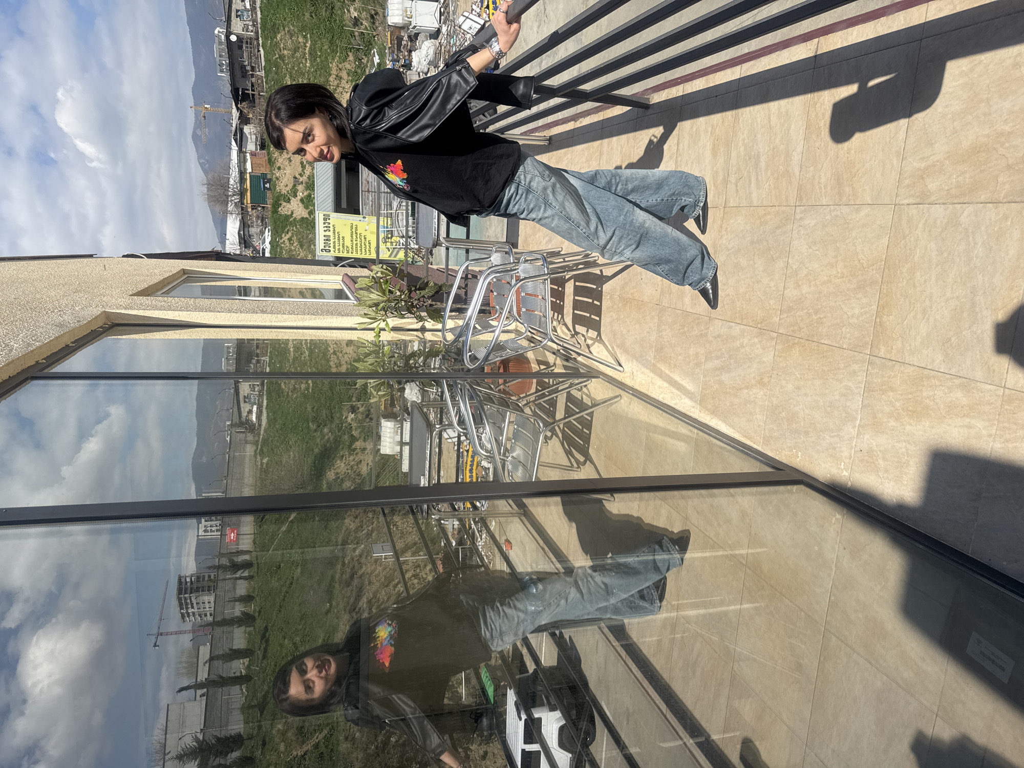 A young adult is photographed on a balcony in Tbilisi, Georgia, with a view of the city. The individual, dressed in a black leather jacket, a dark t-shirt with a colorful design, and light blue jeans, is gripping a black metal railing. The balcony floor is tiled in a light brown color, and there are several metal chairs and a small table with plants nearby. The building has large windows reflecting the outside environment. In the background, the cityscape of Tbilisi is visible under a cloudy sky.