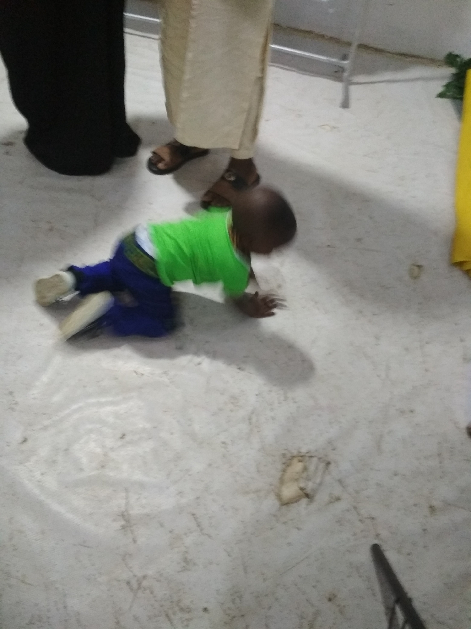 A baby crawls on a white floor, wearing a green shirt and blue pants. Two adults stand nearby, one in black clothing and the other in beige. The location is unknown, but the floor is white.
