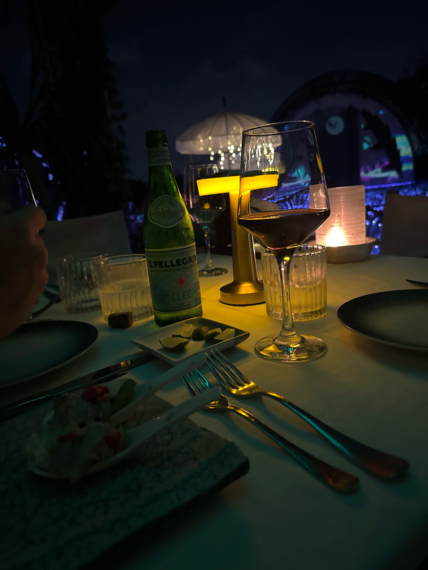 The image captures a dimly lit table setting, likely at a restaurant in North Kuta, Indonesia. A bottle of San Pellegrino sparkling water stands prominently on the table, next to a glass of red wine. Cutlery and plates are arranged for a meal, with a small appetizer served in a white dish. In the background, soft lighting from candles and decorative elements creates an intimate ambiance. The scene suggests a relaxing evening, possibly a dinner date or a meal with friends, in a tropical setting given the presence of palm leaves in the background, indicating the restaurant's location.