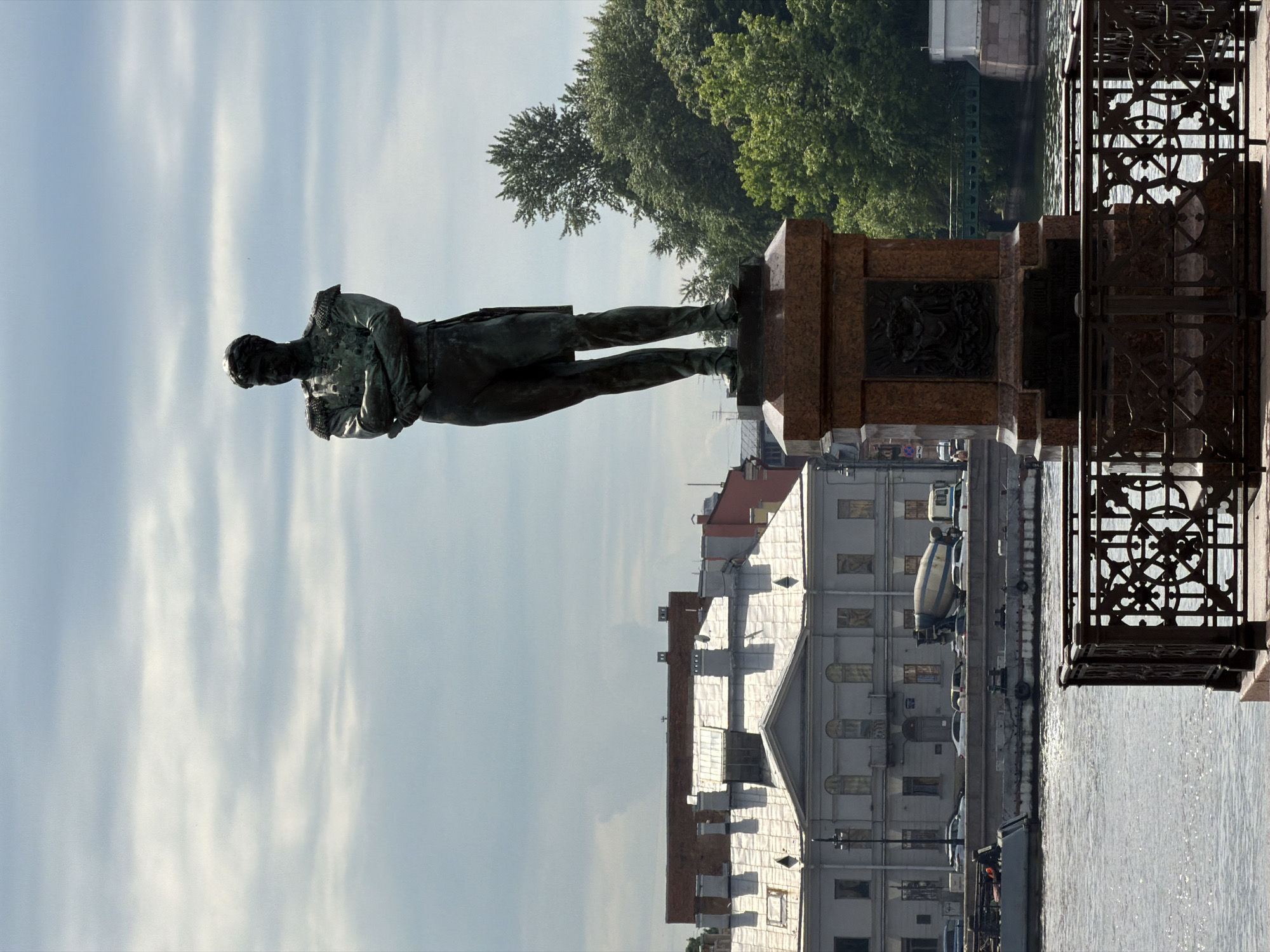 A large bronze statue of a man stands prominently on a reddish-brown granite pedestal, positioned on the left side of the frame. The man is depicted in historical formal wear, including a long coat and boots, with his left arm bent across his body. To the right of the pedestal, an ornate black wrought-iron railing with intricate circular and scroll patterns borders a body of water, likely a river or canal, which glistens under the daylight.
Across the water, on the opposite bank, a line of buildings with varying architectural styles forms the backdrop. One building is particularly noticeable with a light-colored, textured roof and numerous windows. On the street alongside these buildings, several vehicles are parked, including a distinct cement mixer truck. Dense green trees are visible behind the buildings and extend into the midground behind the statue. Above, the sky is light blue with scattered, elongated white clouds.
The image captures a static, daytime urban scene. The bright light and clear skies suggest a sunny day. The calm water surface shows reflections and small ripples. No specific event is taking place. The architectural style of the buildings and the design of the monument are characteristic of a historic European city, fitting with the location context of Saint Petersburg, Russia. There is no discernible text visible in the image.