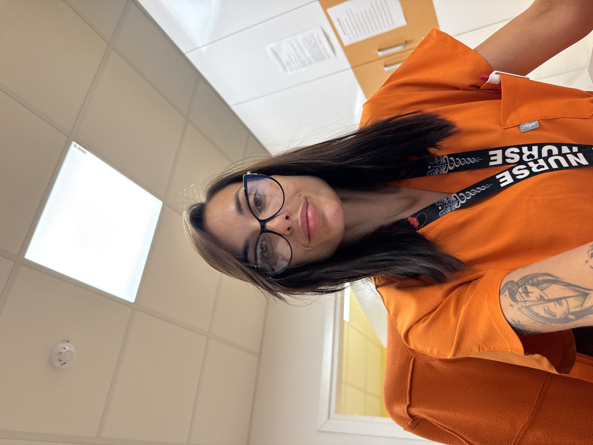 A woman is facing the camera while wearing an orange scrub top with a "Nurse" lanyard around her neck. She has dark hair, wears glasses, and has a tattoo on her arm. The background shows a tiled ceiling with a square light fixture and a fire alarm. There are also filing cabinets and a window visible. There is no indication of the city of Pleven, Bulgaria.