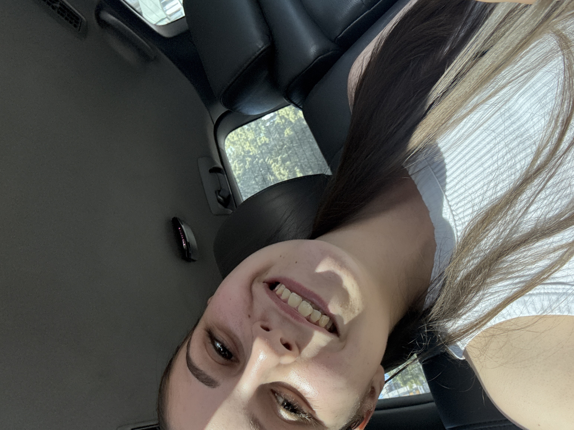 The image captures a young woman inside a car, presented at an inverted angle, taking a selfie or being photographed. She is smiling broadly and looking directly at the camera, conveying happiness or joy. Her hair is long, a mix of dark roots and lighter, possibly sun-bleached, brown ends. She is wearing a light-colored, ribbed top.
The setting is clearly a car interior, visible through details like dark-colored seats and headrests, an overhead light fixture, and air vents on the ceiling. Through the car's rear window, green foliage, indicative of trees or bushes, can be seen, suggesting an outdoor location. Bright natural sunlight illuminates the scene, casting shadows on the woman's face and highlighting her features, which points to a sunny daytime setting. No specific activities beyond posing for the photo are evident, and there is no visible text in the image. The geographical context for the image is Burgas, Bulgaria.